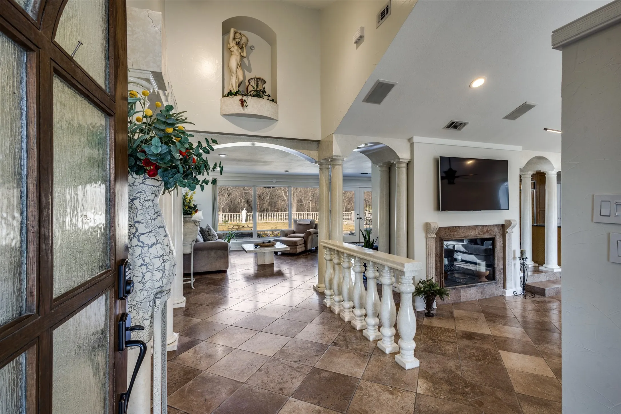 Open foyer view showcasing walnut travertine floors, marble columns, and balustrade accents. Floor-to-ceiling windows at the rear bring in natural light and frame wooded views. Central fireplace anchors the main living area and connects the primary gathering spaces.