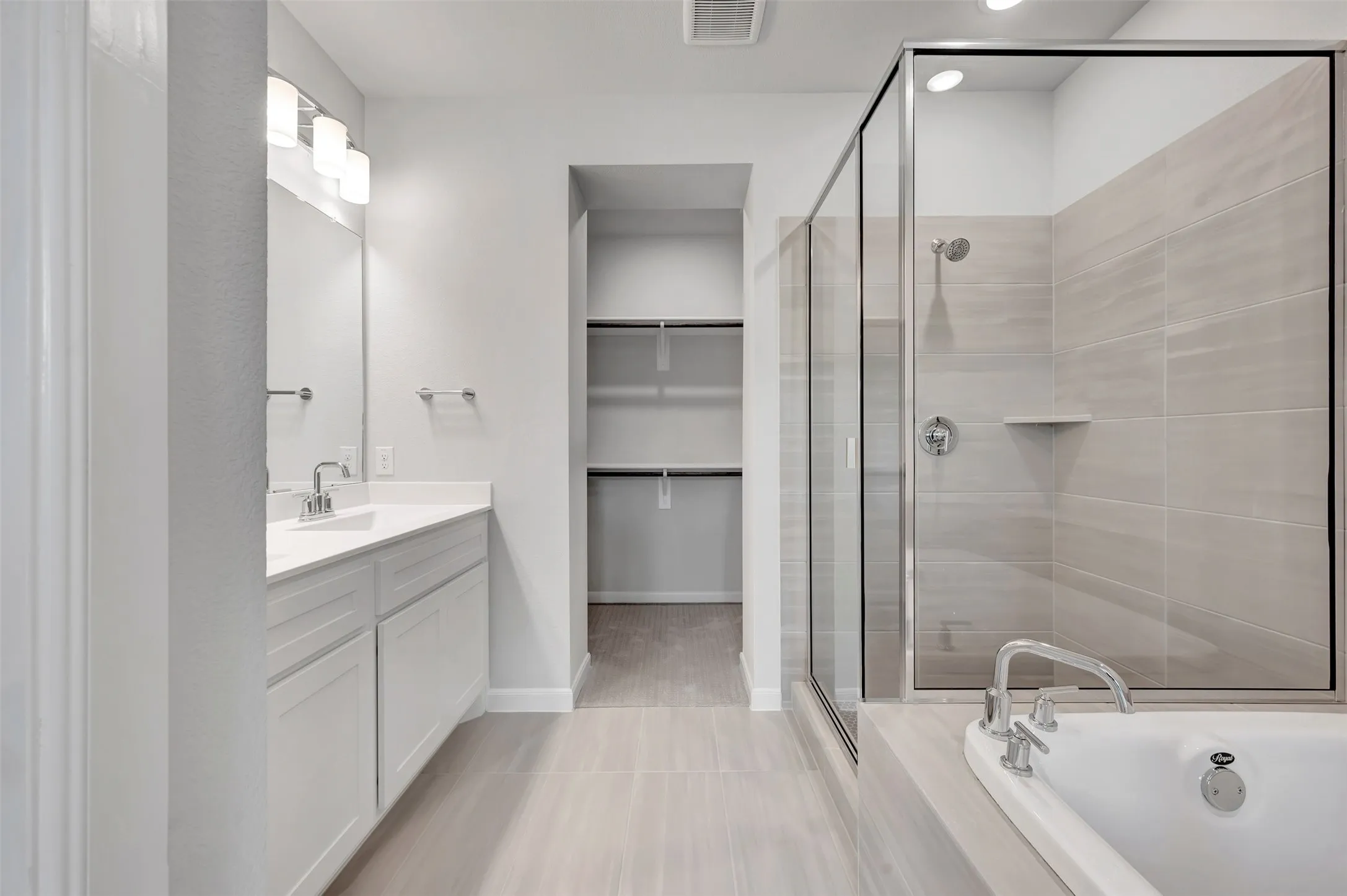 Bathroom featuring a dual vanity with white cabinetry and a large mirror, a glass-enclosed shower with tiled walls and a built-in niche, a soaking tub with chrome fixtures, and a walk-in closet with built-in shelving