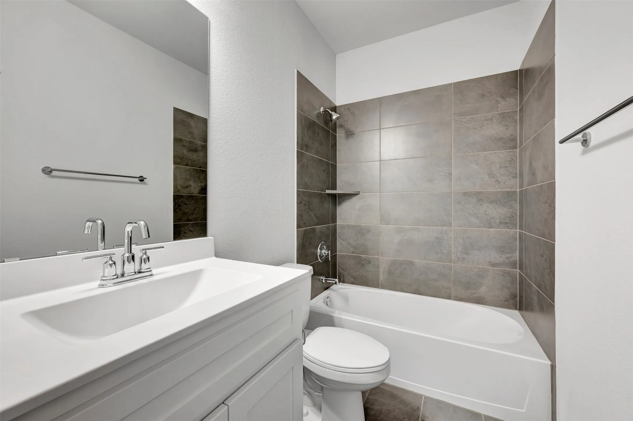 Bathroom featuring a single white vanity with a rectangular sink and chrome faucet, a large wall-mounted mirror, a tub/shower combination with large format gray tile surround, an integrated shower shelf, and a chrome towel bar