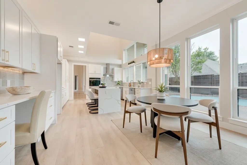This bright kitchen and dining area features extensive white cabinetry with gold hardware, light wood flooring, and a dining table with four chairs illuminated by a contemporary pendant light