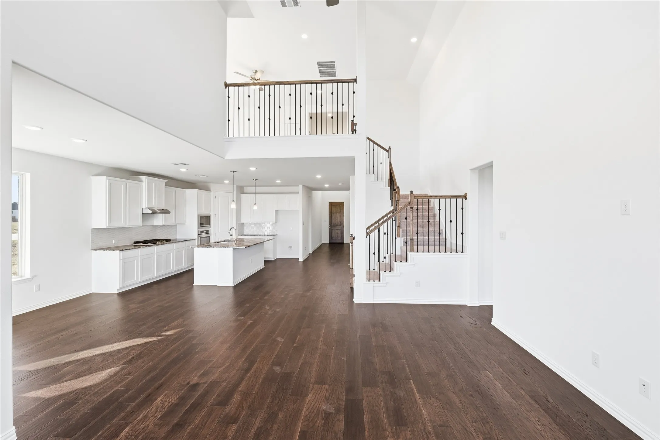 Expansive open-concept interior featuring high ceilings, wood-finish flooring, a contemporary kitchen with white cabinetry and an island, and a prominent staircase with wrought iron balusters