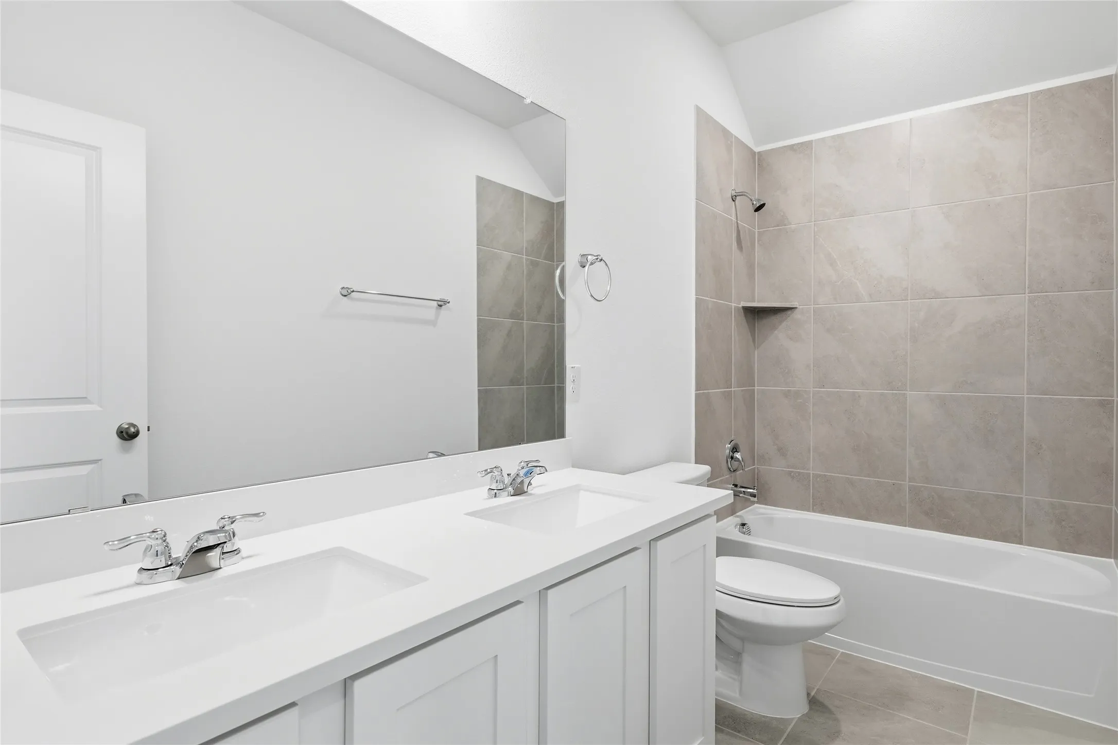 Bathroom featuring a dual vanity with white cabinetry and integrated sinks, chrome fixtures, a full-width wall mirror, a bathtub with a tiled surround, and floor-level tiling