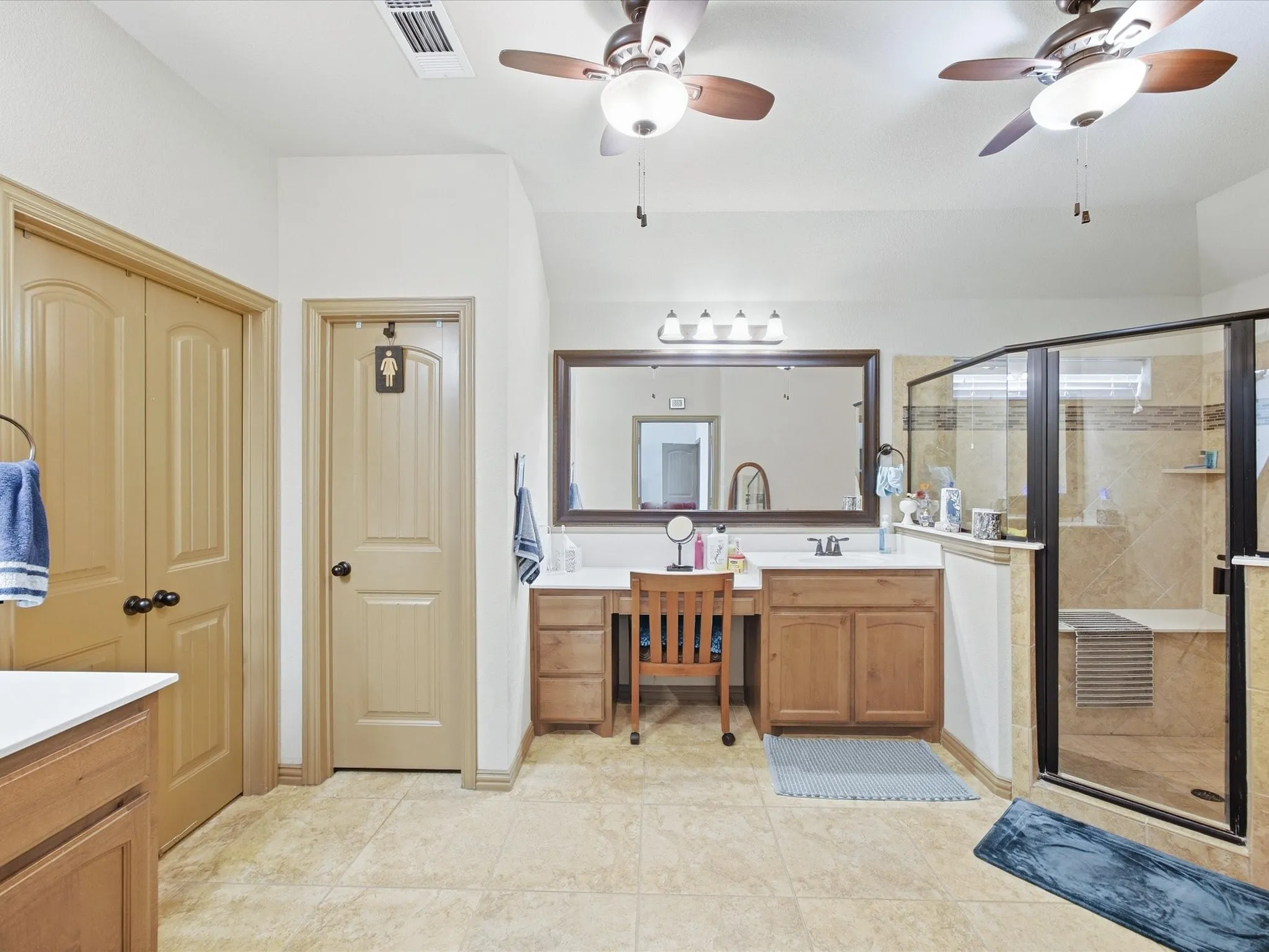 Dual ceiling fans, natural wood finish vanity with integrated sink, large framed mirror, vanity lighting fixture, and a glass-enclosed shower with tiled surround