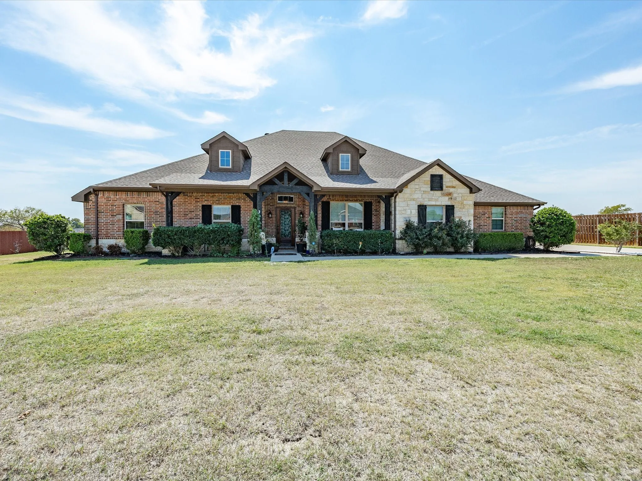 Brick and stone exterior with dormer windows, covered front entry, dark wood accents, and mature landscaping