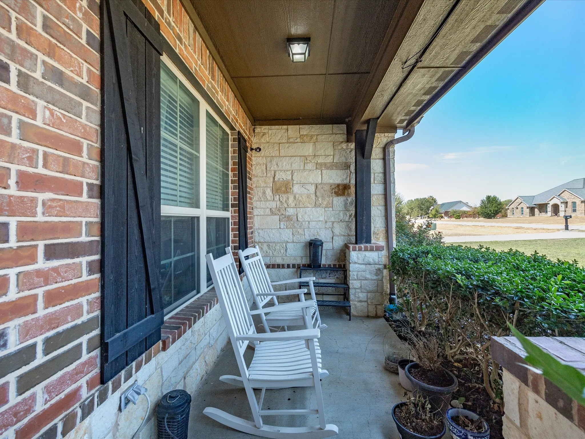 Covered front porch featuring a brick and stone exterior, dark wood shutters, and an overhead light fixture