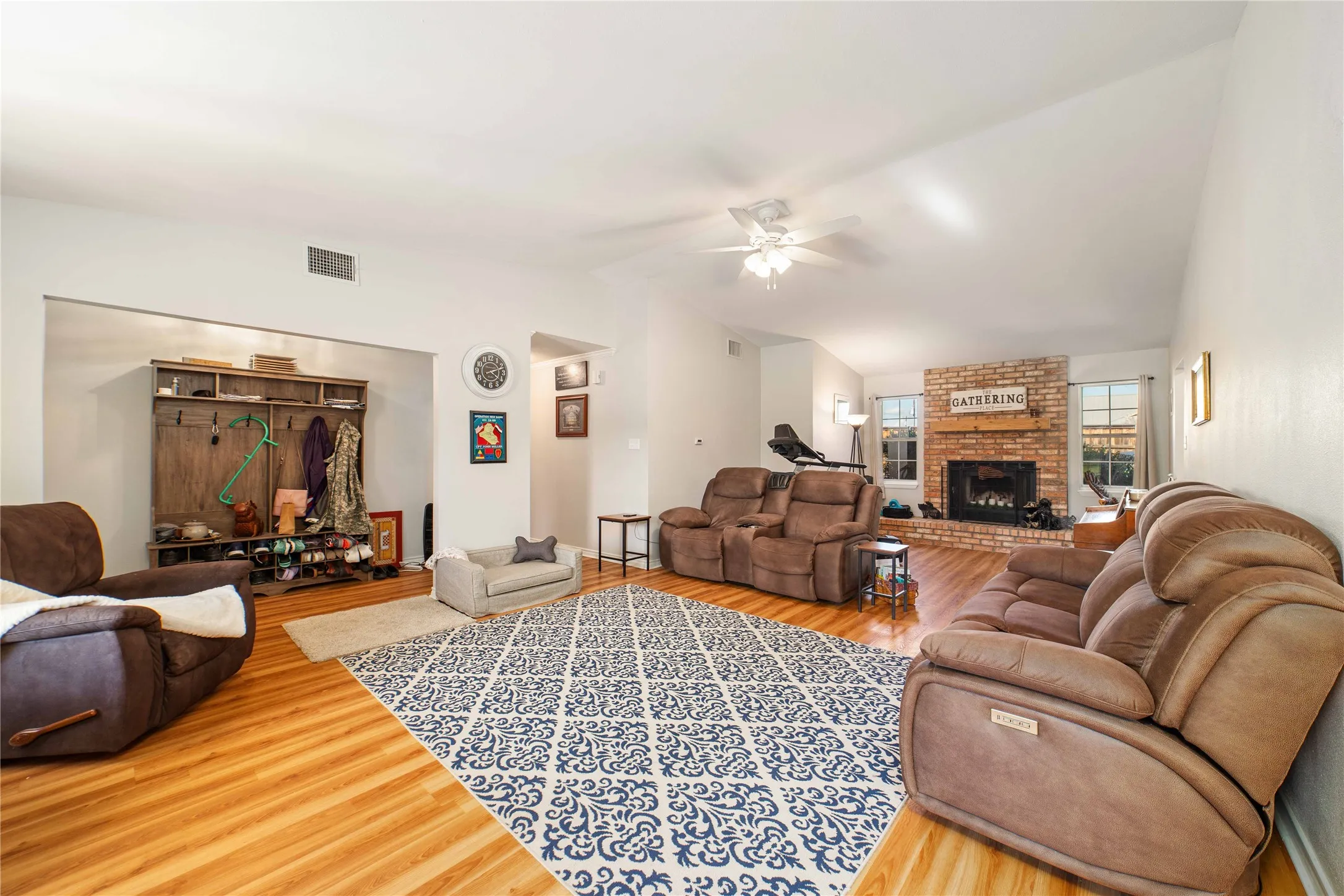 Living room featuring vaulted ceiling, a ceiling fan, wood finished floors, a brick fireplace, and visible vents