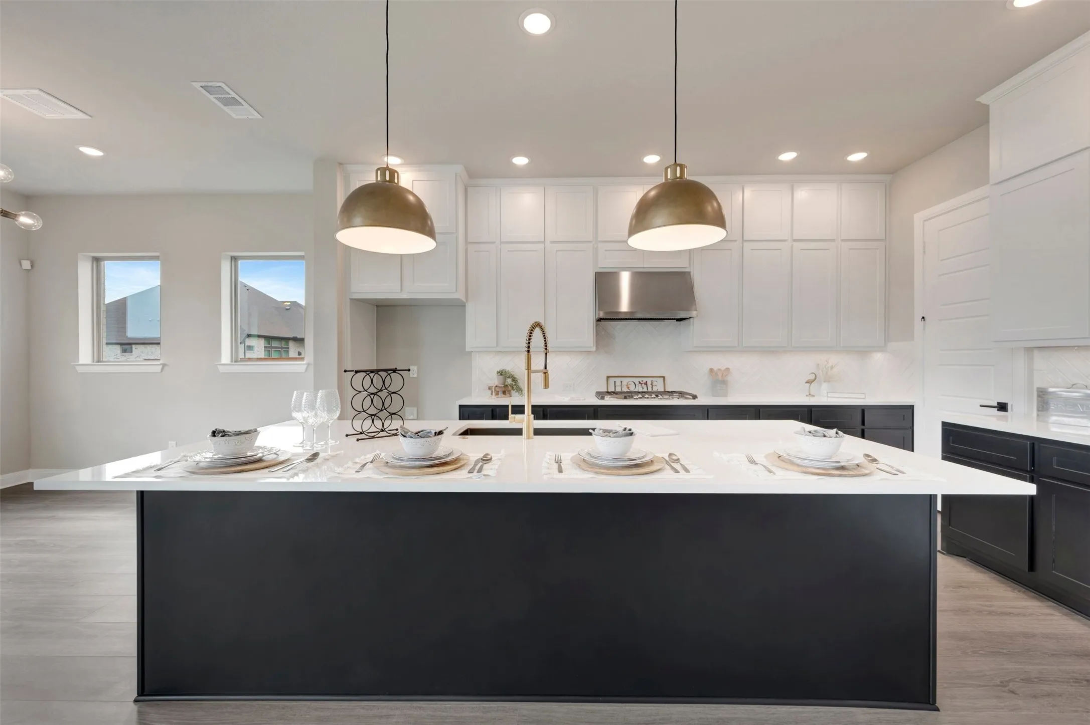Spacious kitchen featuring a large central island with a white countertop, two brass pendant lights, stainless steel range hood, and wood-finish flooring