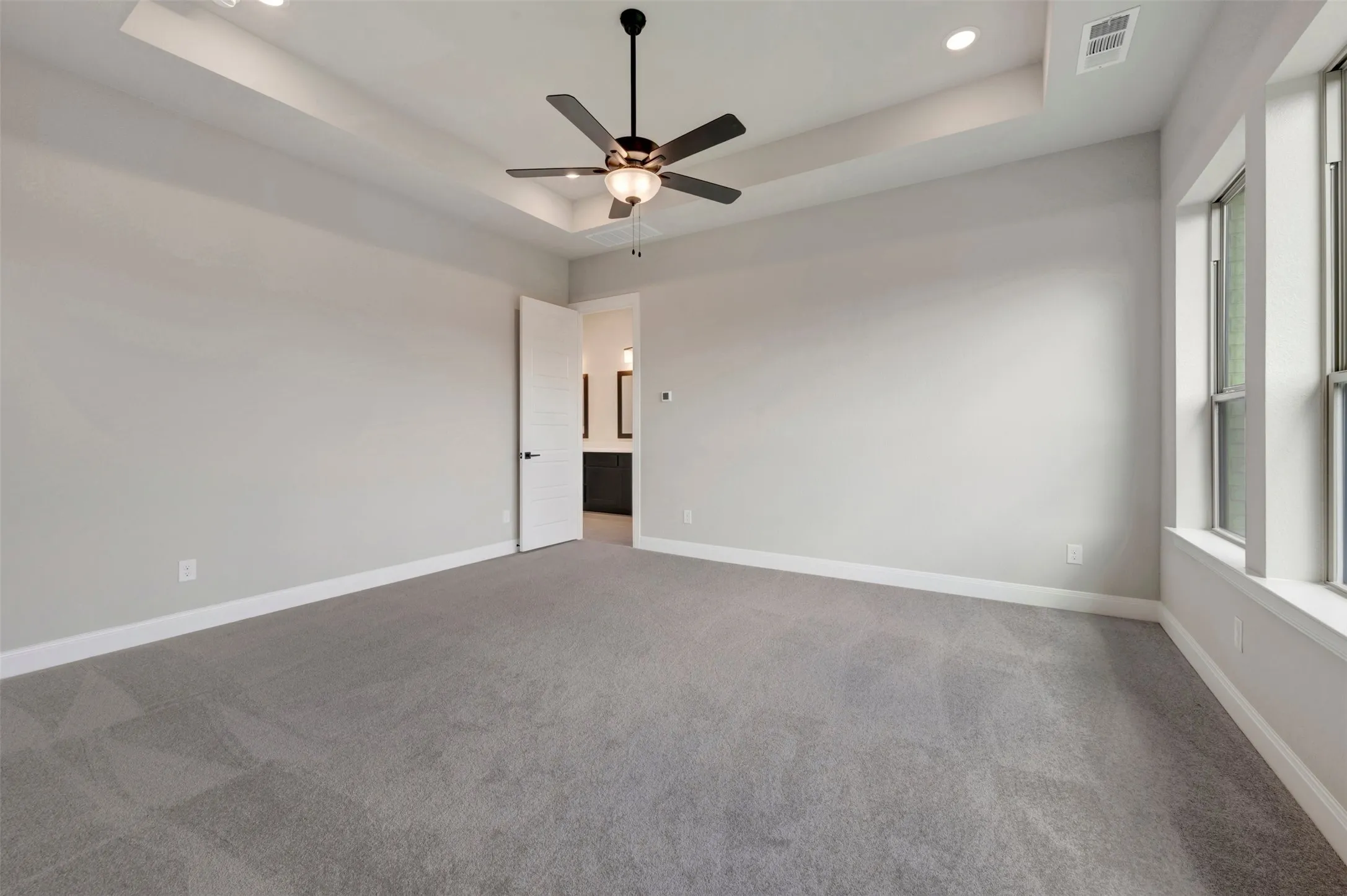 Spacious room featuring tray ceiling, recessed lighting, ceiling fan, plush gray carpeting, and multiple windows