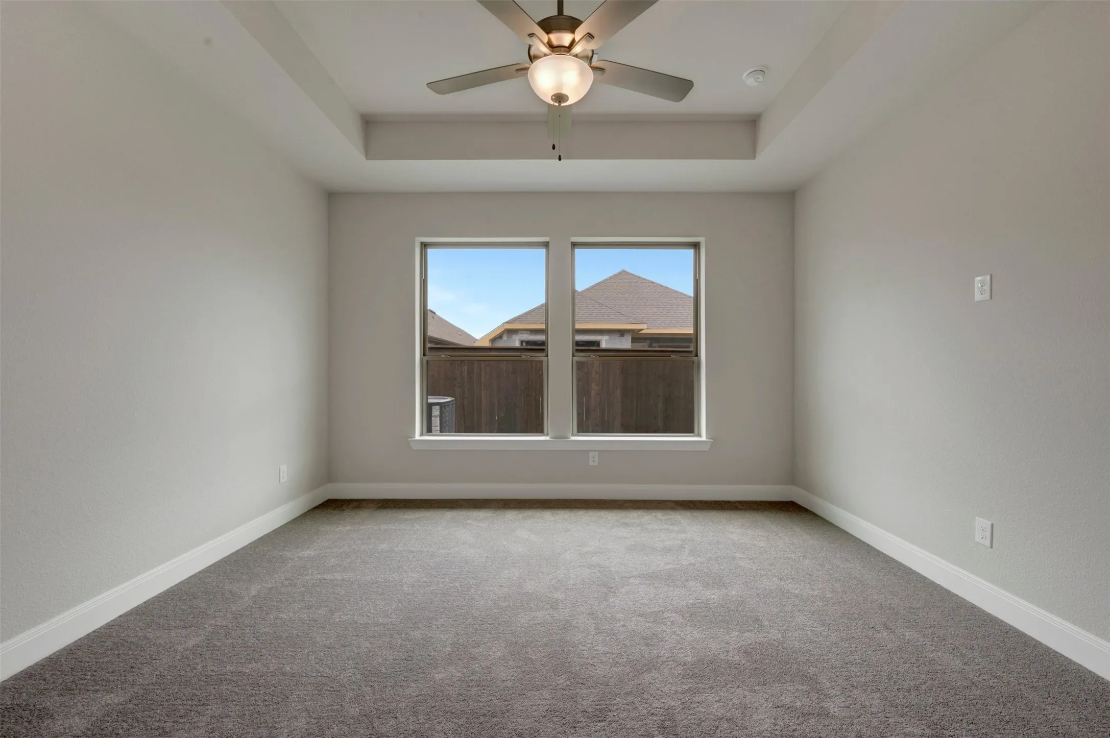 Spacious room featuring a tray ceiling, ceiling fan with integrated lighting, twin windows, neutral wall tones, and gray carpeting