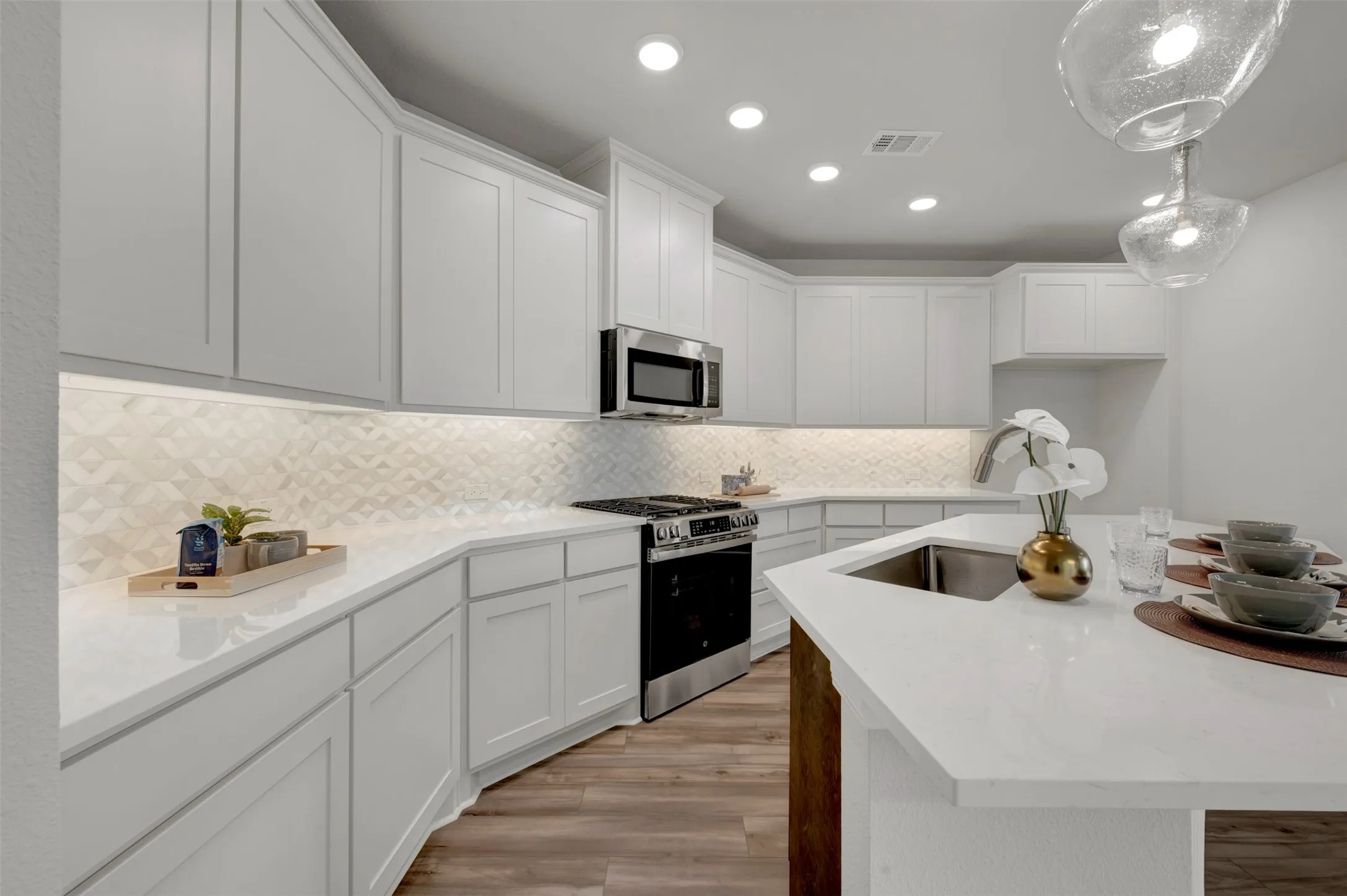 Kitchen featuring white shaker-style cabinetry, white countertops, textured tile backsplash with under-cabinet lighting, stainless steel appliances, and a central island with an undermount sink