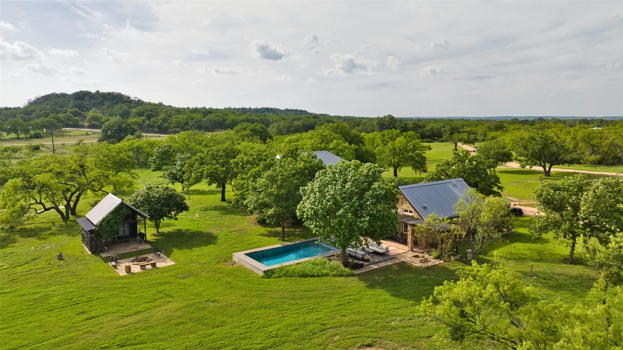 Expansive property featuring a main residence with a metal roof, a rectangular swimming pool with an adjacent patio, and a separate structure with a covered porch and outdoor fire pit area