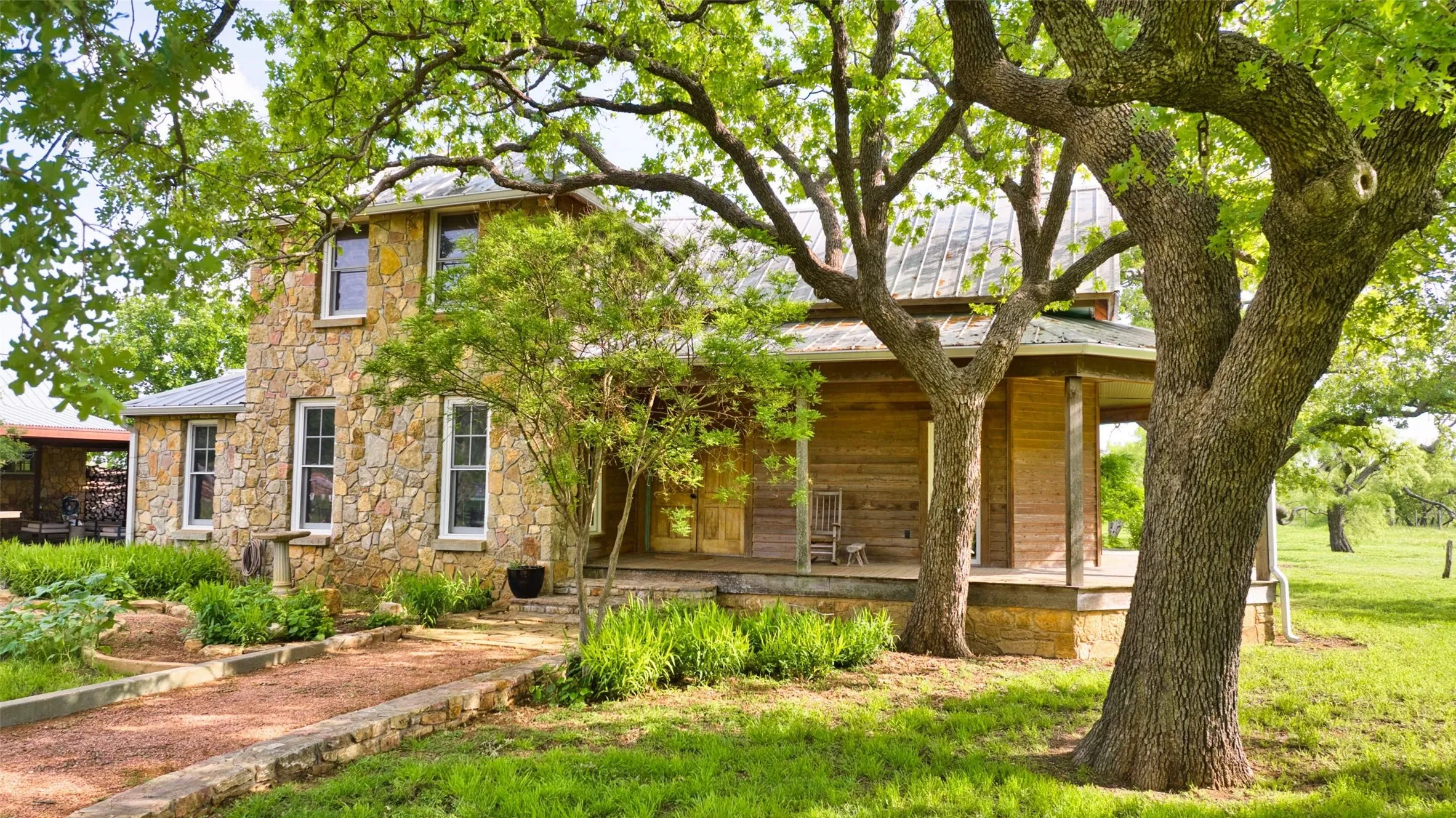Two-story stone and wood-sided residence featuring a wraparound porch, mature trees, a gravel pathway, and lush landscaping