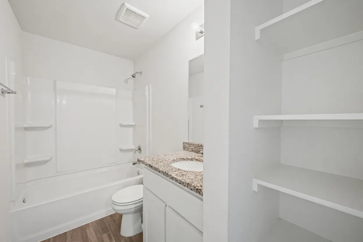 White tub-shower combination, vanity with granite-finish countertop, wood-finish flooring, and built-in shelving