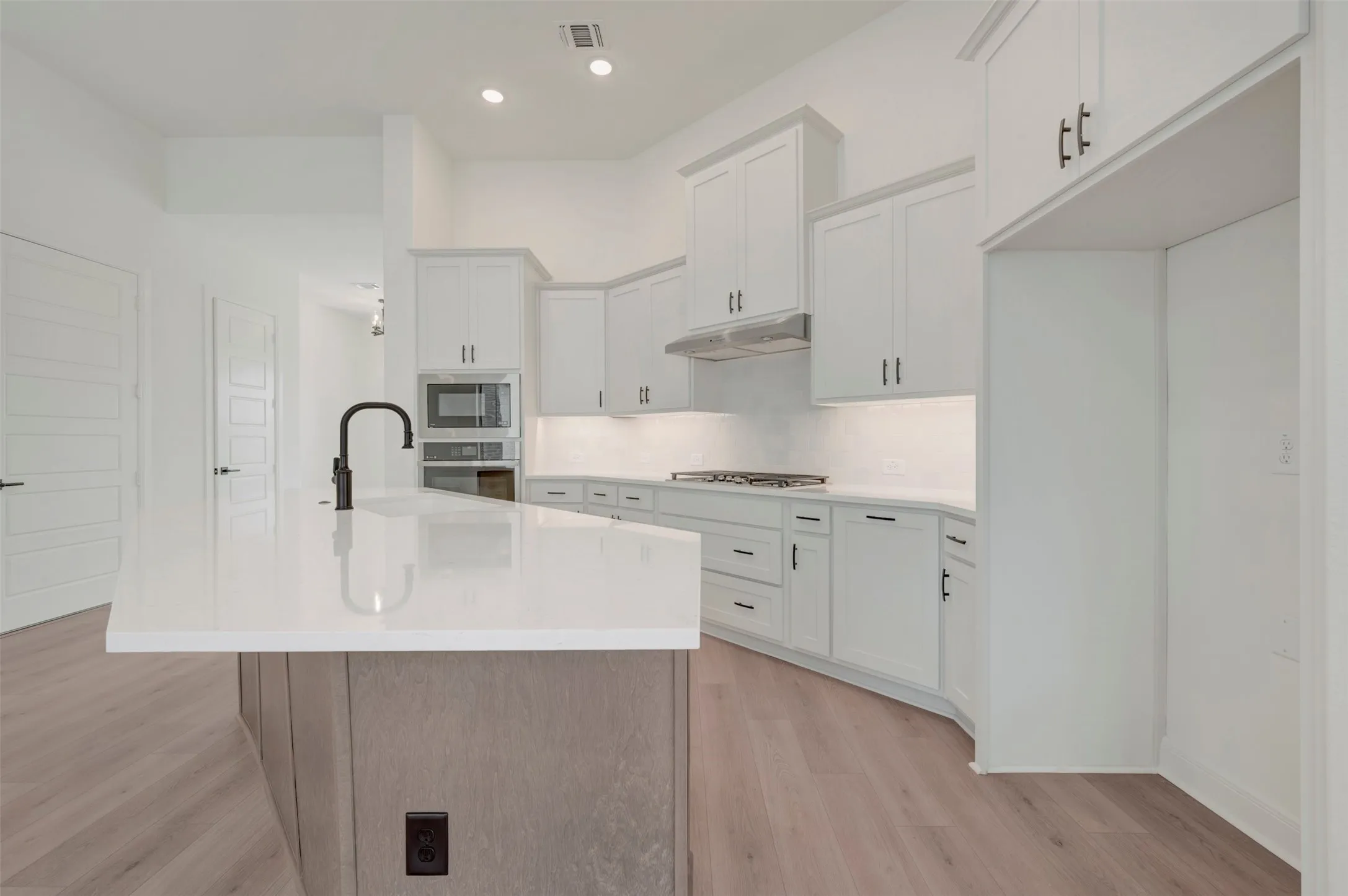 Spacious kitchen featuring a large island with a black gooseneck faucet, white shaker cabinetry, integrated stainless steel appliances, recessed ceiling lighting, and wood-finish flooring