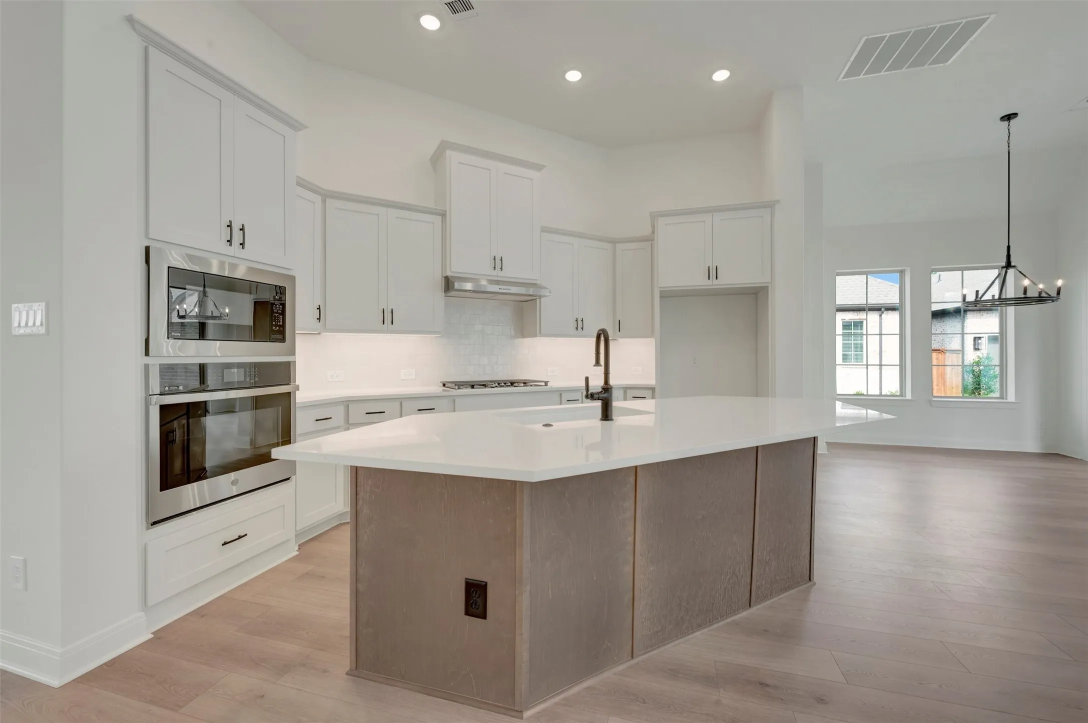 Modern kitchen featuring a large central island with a solid surface countertop, light wood-finish flooring, white cabinetry with dark hardware, stainless steel built-in appliances, and a tiled backsplash