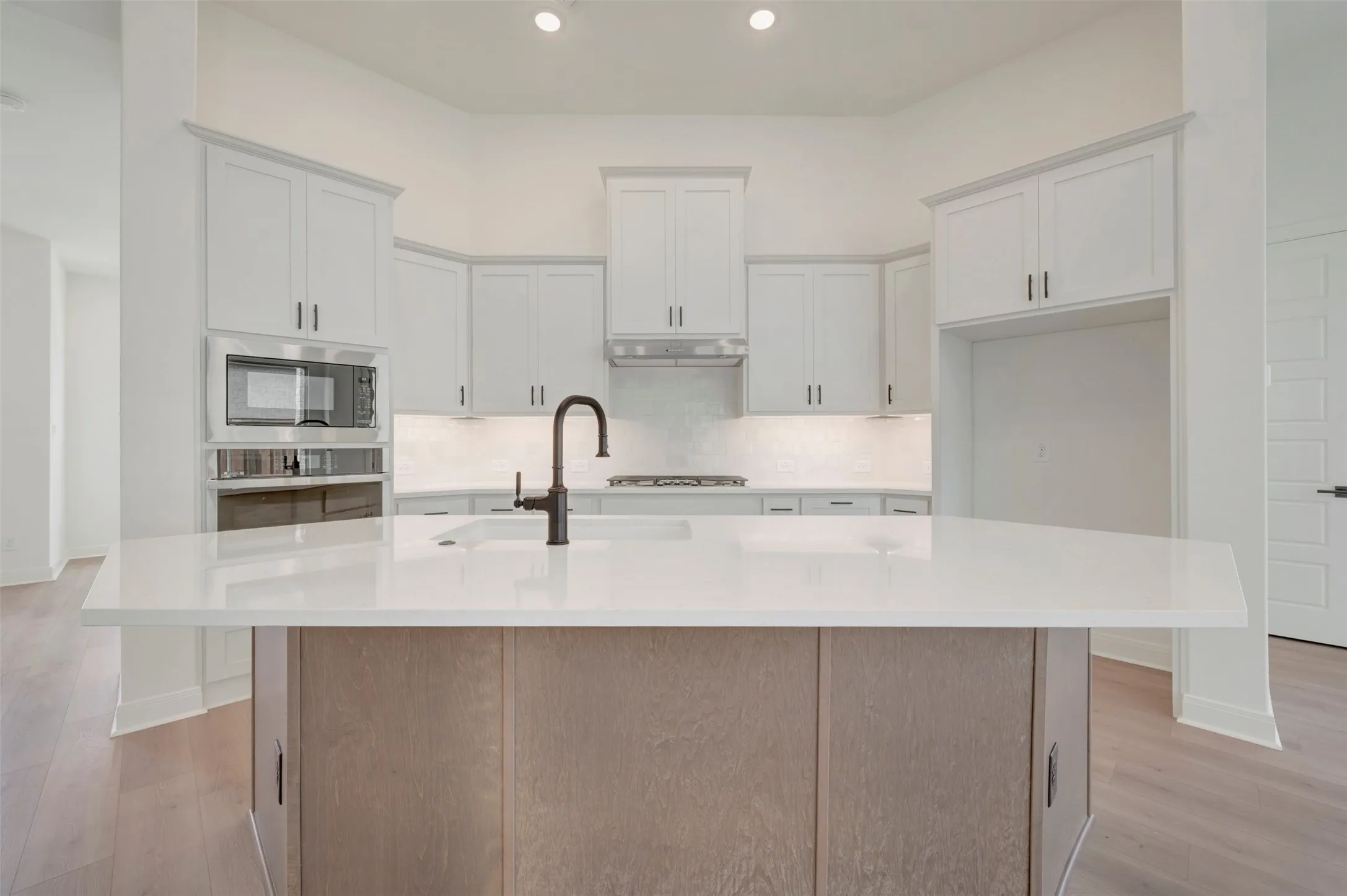 Spacious kitchen featuring a central island with a built-in sink and matte black faucet, light wood-finish cabinetry, white countertops, built-in microwave, and recessed lighting