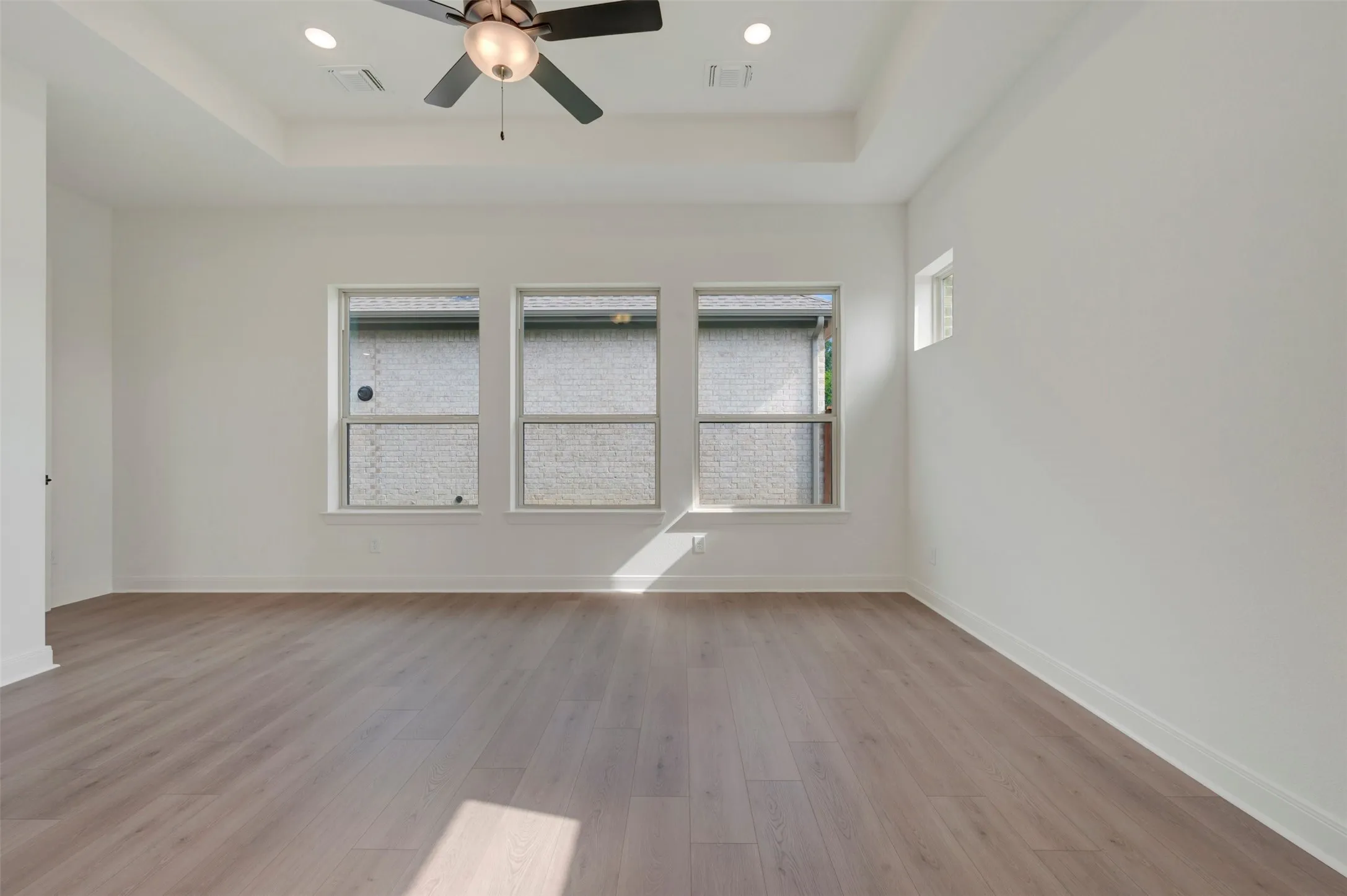 Spacious room featuring light wood-finish flooring, a tray ceiling with recessed lighting, and a contemporary ceiling fan