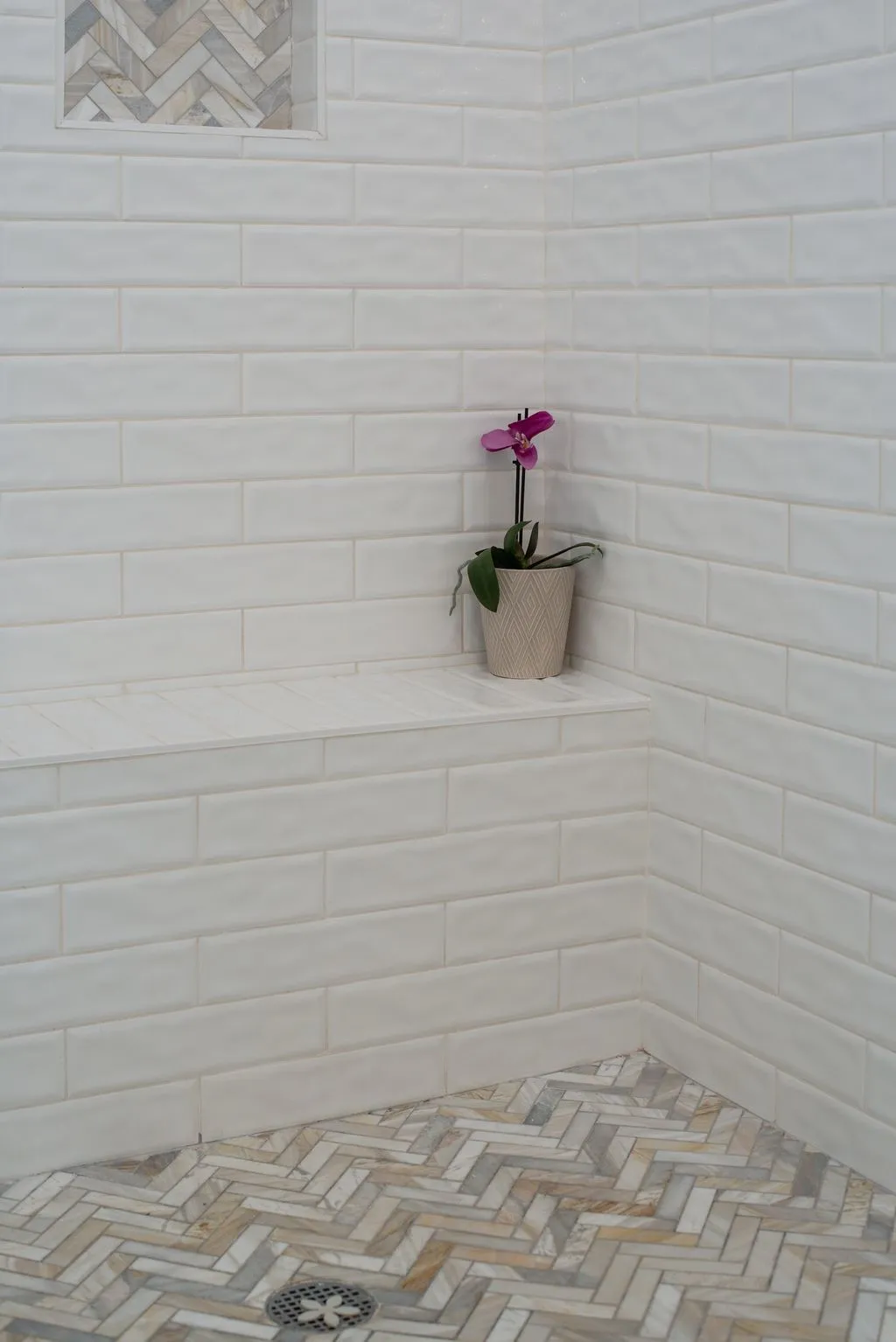 White subway tile walls, built-in shower bench, recessed niche with herringbone tile, and herringbone tile floor