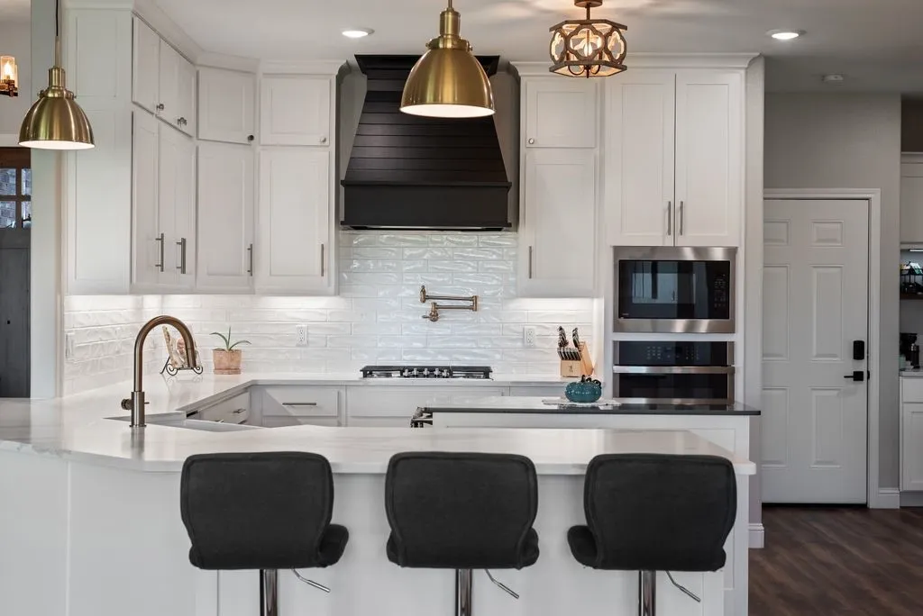 White cabinetry with brushed nickel hardware, stainless steel built-in microwave and oven, a white textured tile backsplash, a curved kitchen island with a built-in sink, and a dark wood range hood
