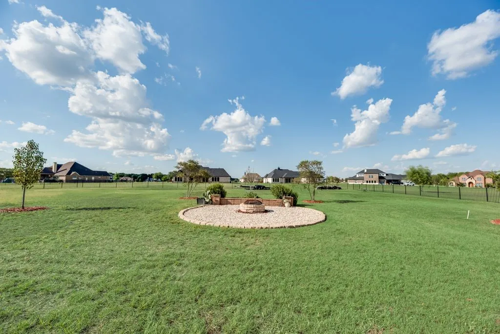 Expansive green space featuring a circular stone patio with a built-in fire pit, surrounded by a metal fence