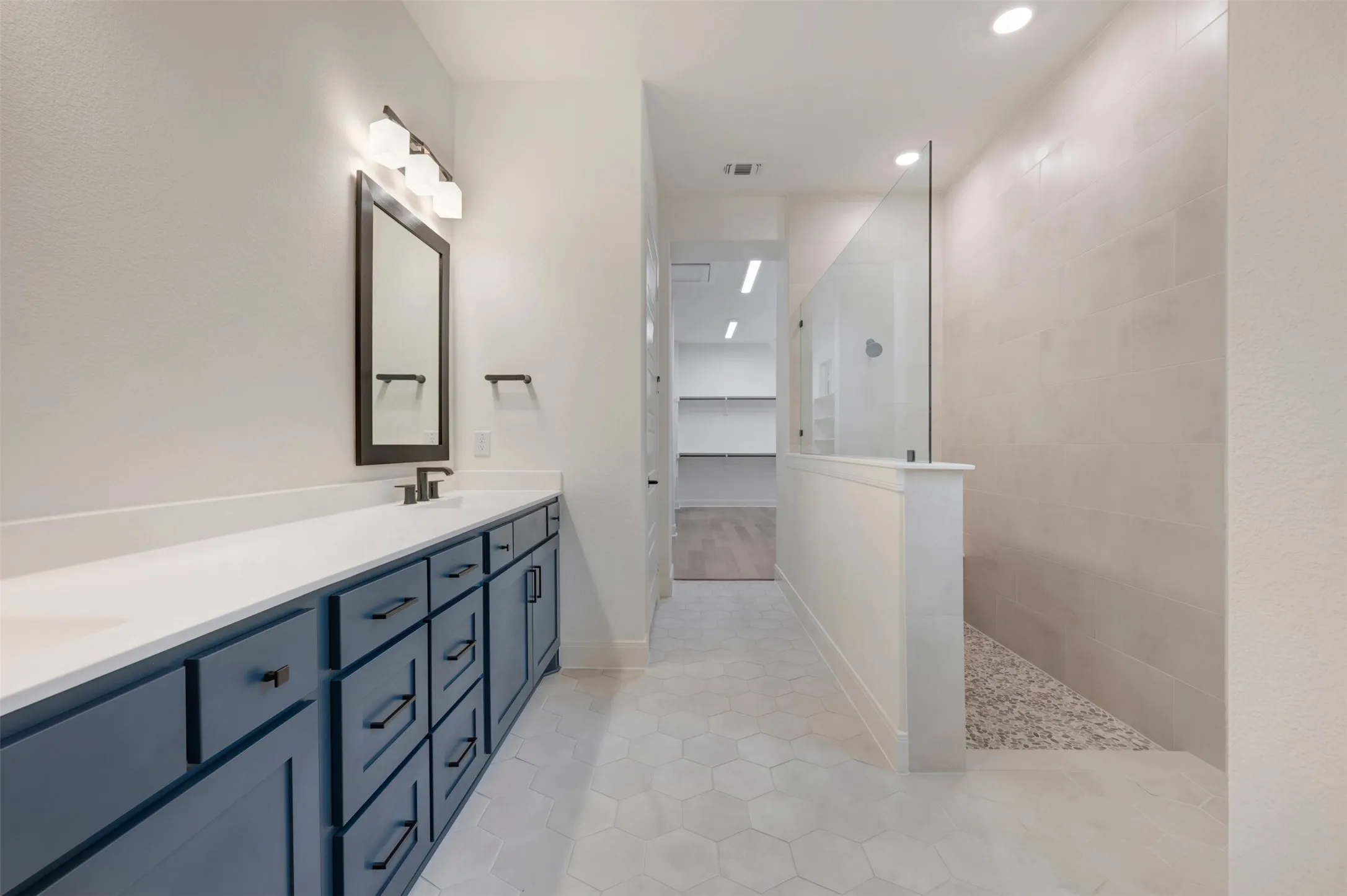Bathroom featuring hexagon tiled floors, a long vanity with blue cabinetry and white countertops, a mirror with sconce lighting, and a glass-enclosed shower with pebble flooring
