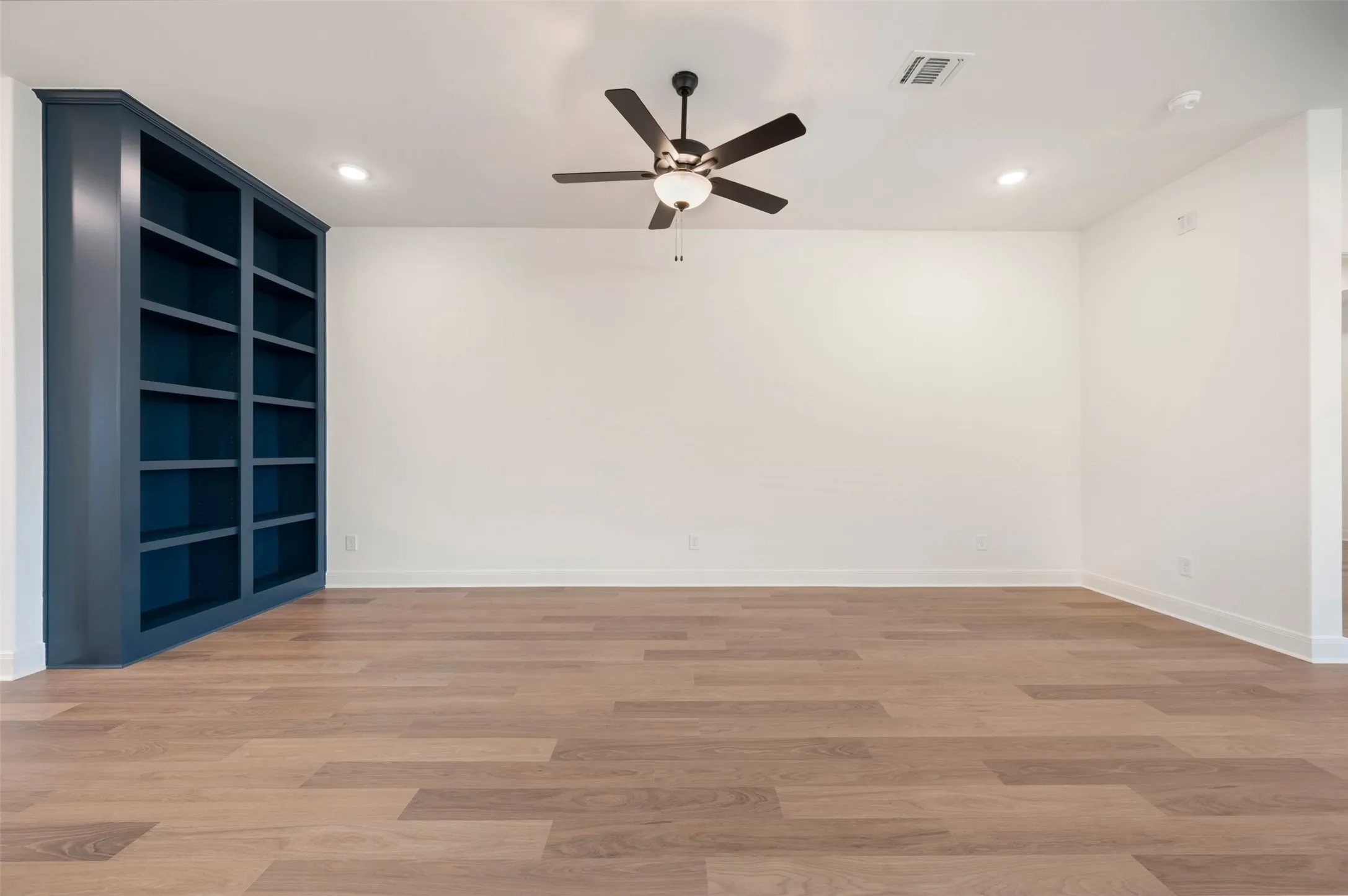 The room features light-colored flooring, a ceiling fan with integrated lighting, recessed lighting, and a built-in dark blue bookshelf
