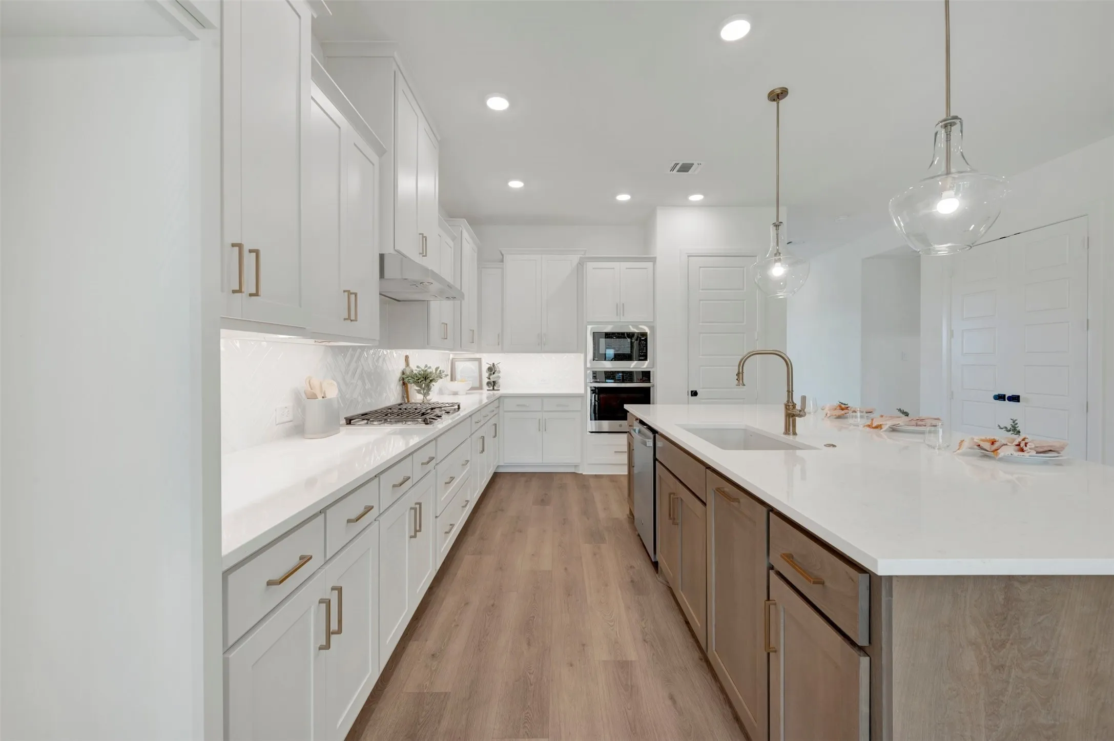 Modern kitchen featuring white shaker cabinetry with gold-tone hardware, a light-toned wood-finish flooring, a central island with an undermount sink and gooseneck faucet, and recessed lighting with two glass pendant fixtures