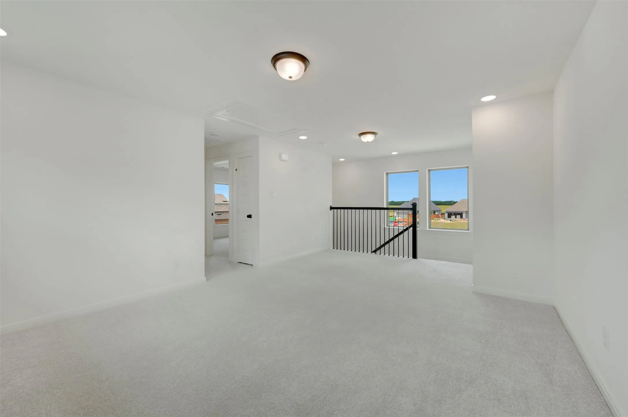 Spacious loft area featuring light gray carpeting, crisp white walls, multiple windows, recessed lighting, and a black metal spindle railing