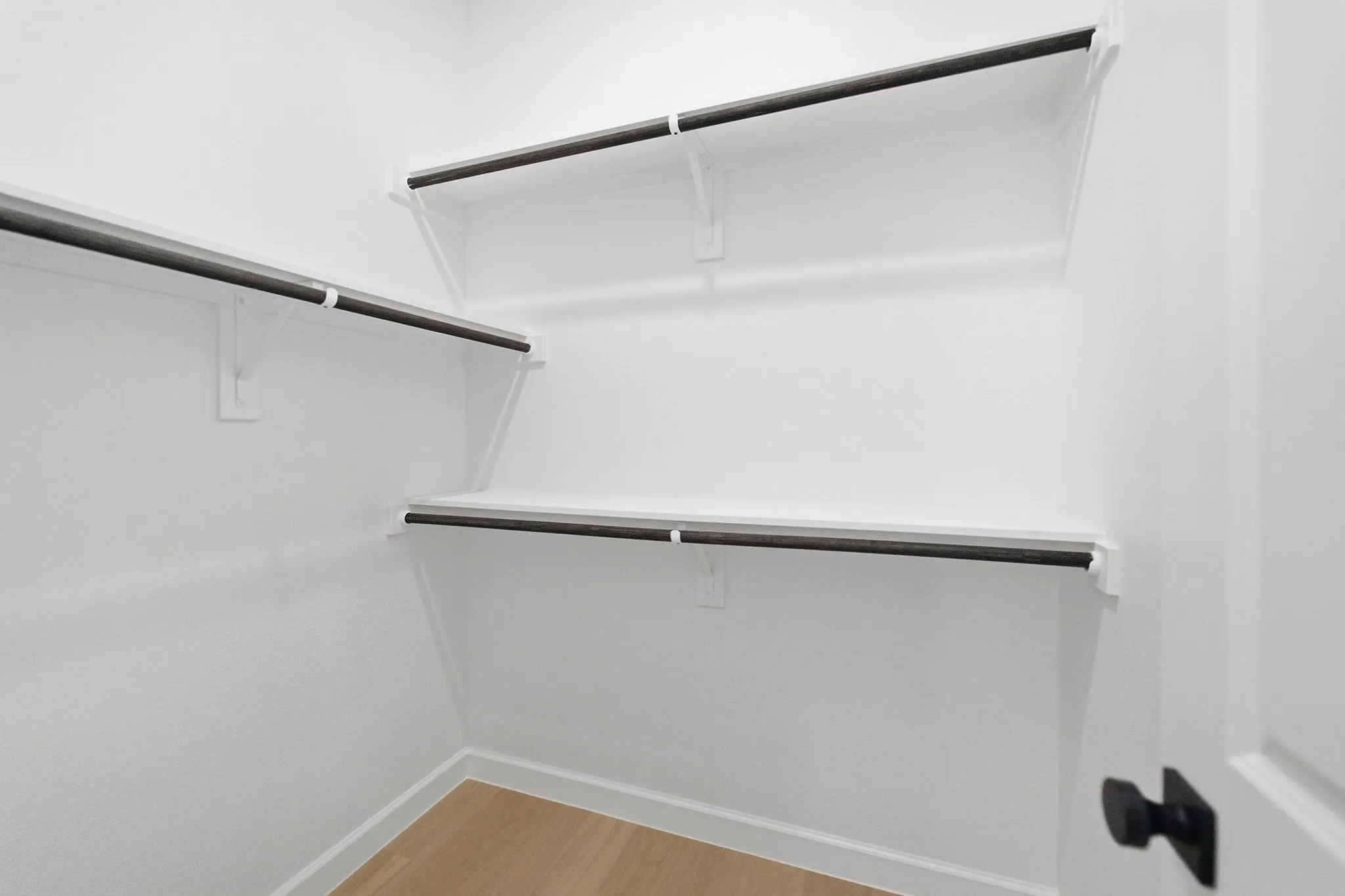 White walk-in closet featuring light wood-finish flooring, white walls, and multiple built-in shelving units with black hanging rods