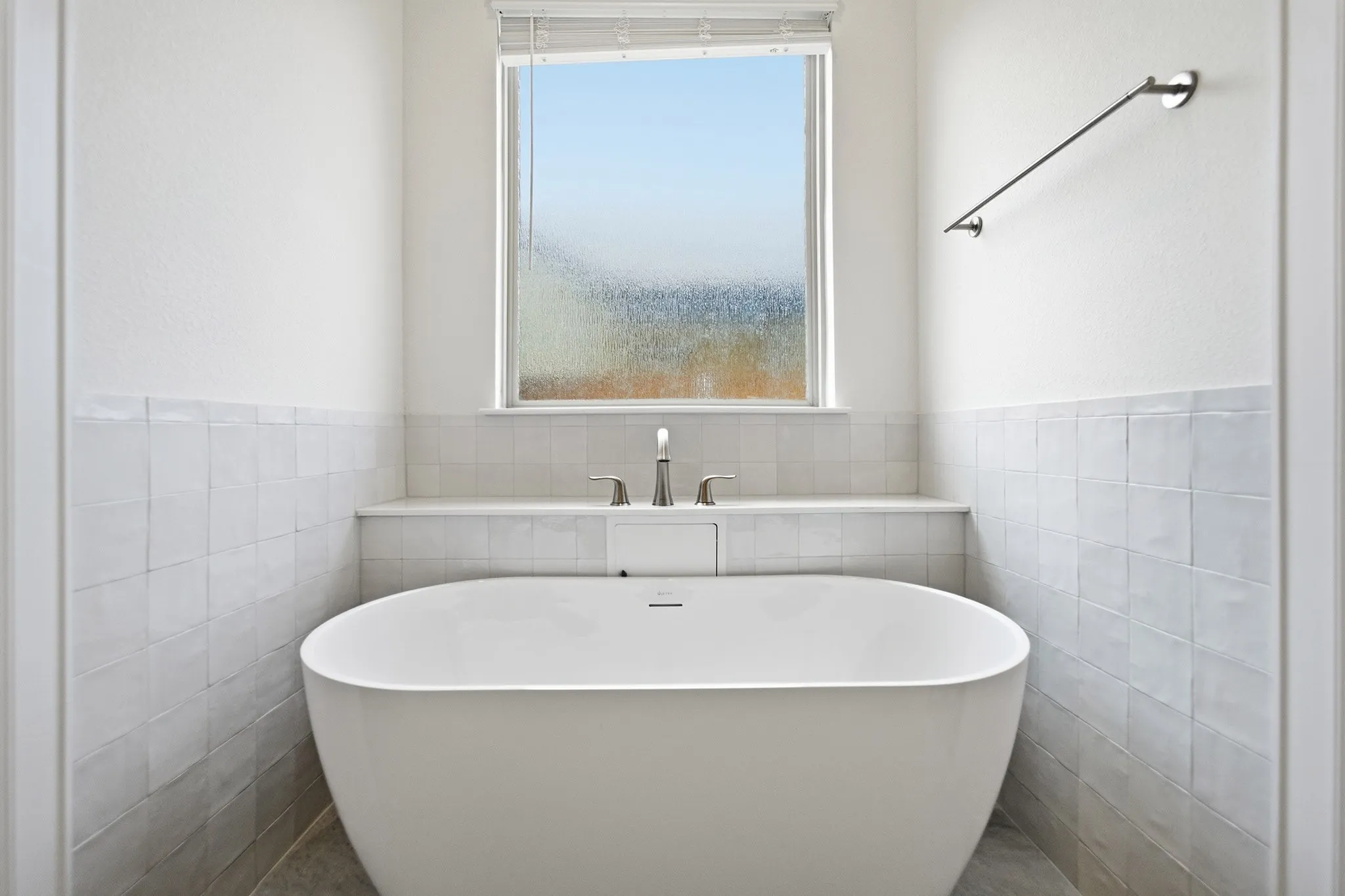 Freestanding soaking tub with polished chrome fixtures, surrounded by white square wall tiles and a built-in ledge