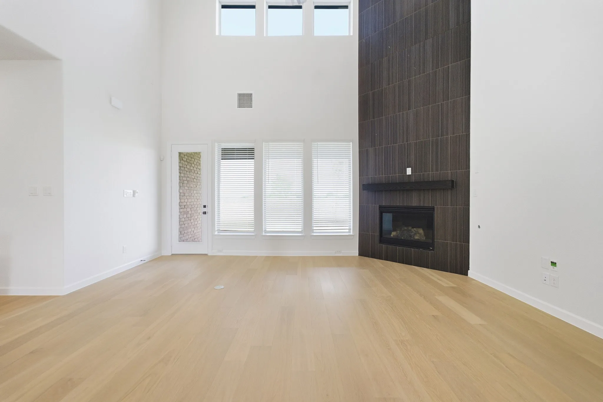 High-ceiling room featuring light wood-finish flooring and a contemporary fireplace with a dark tile surround and integrated mantle