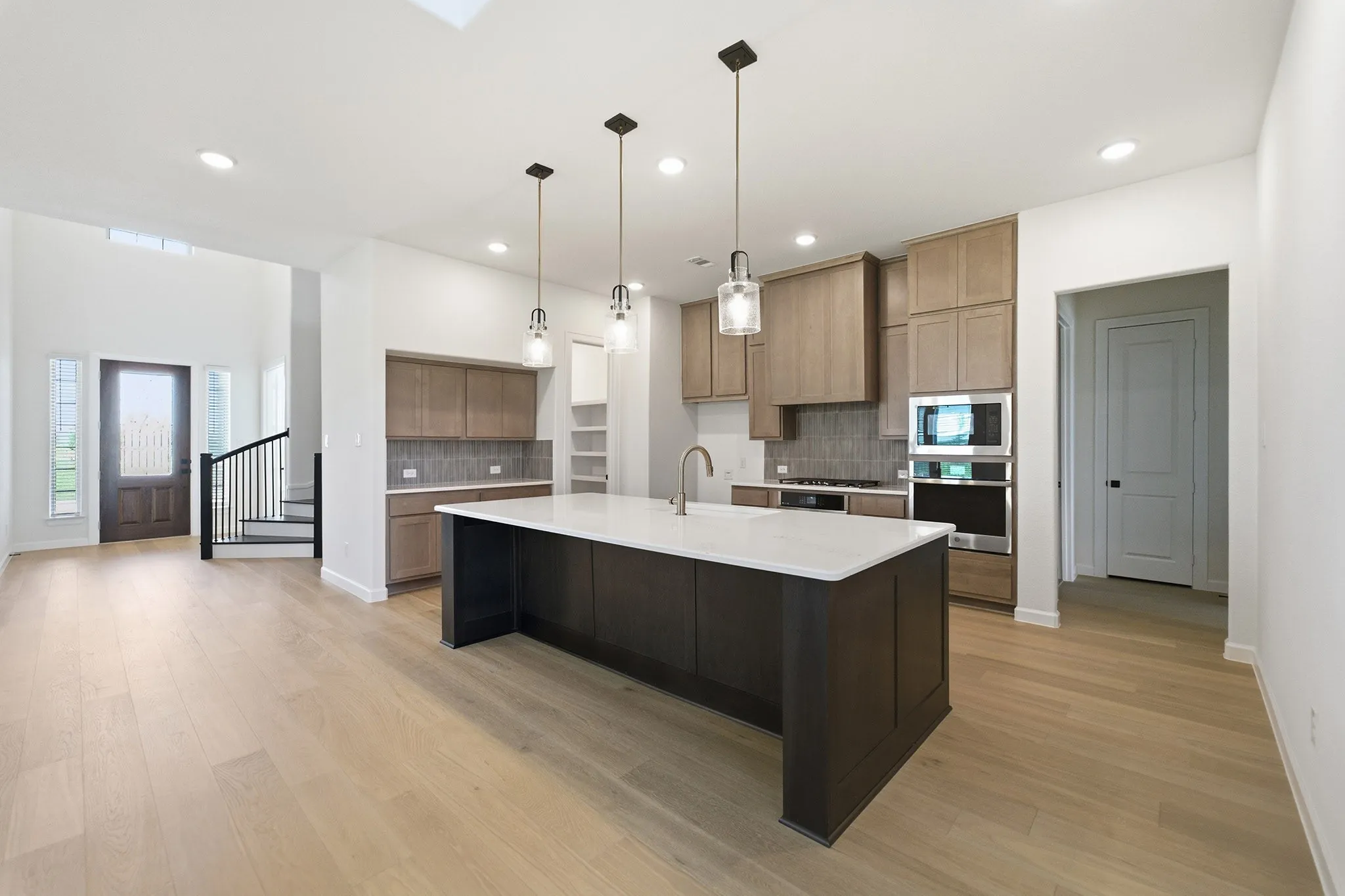 Open-concept kitchen featuring an oversized island with a white solid surface countertop, stainless steel appliances, light wood-finish cabinetry, and wood-finish flooring​​‌​​​​‌​​‌‌​‌‌​​​‌‌​‌​‌​‌​​​‌​​ throughout