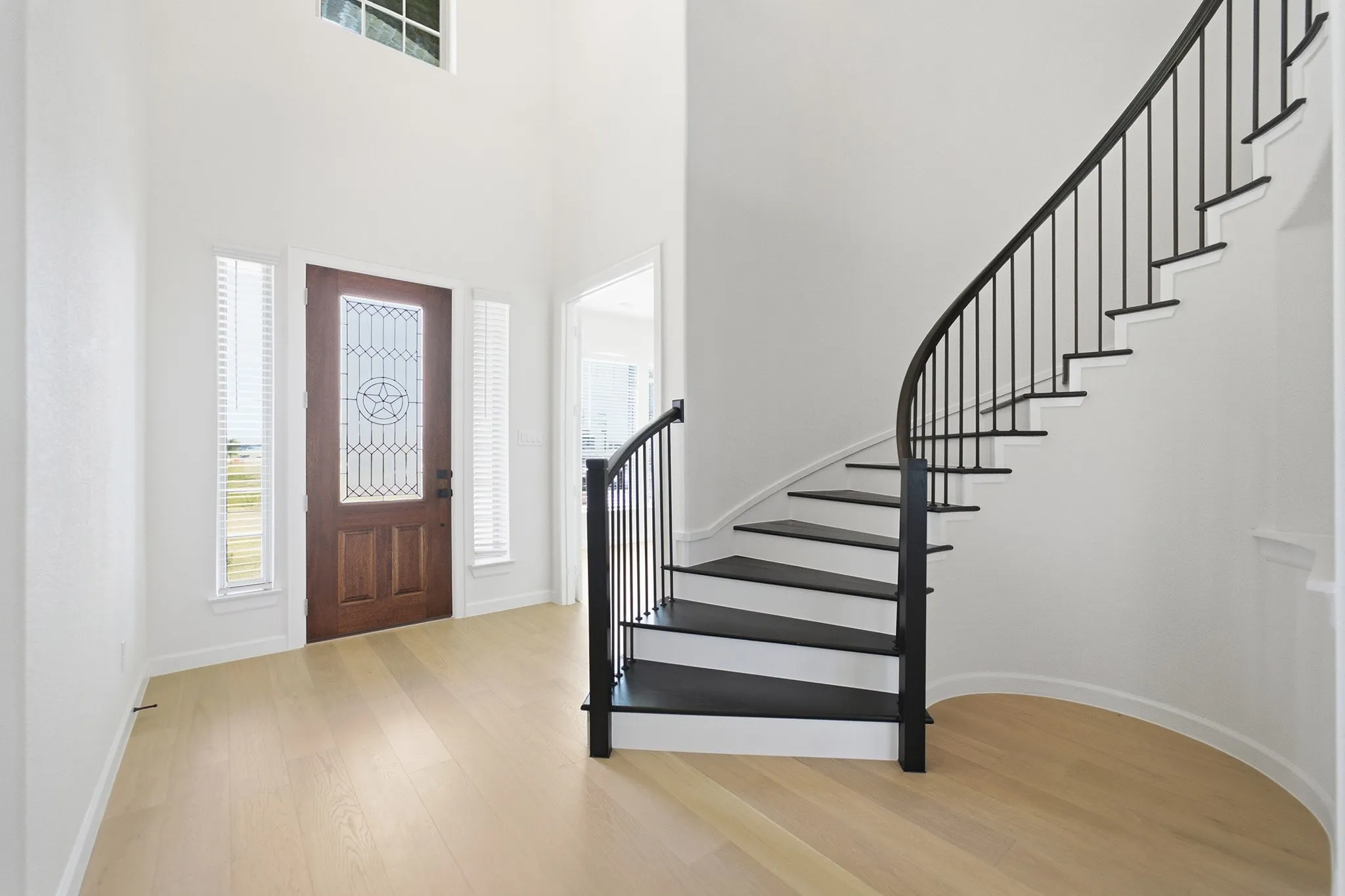 Grand foyer featuring a curved staircase with black metal balusters, a decorative wood-panel entry door with leaded glass, tall windows, and light wood-finish flooring