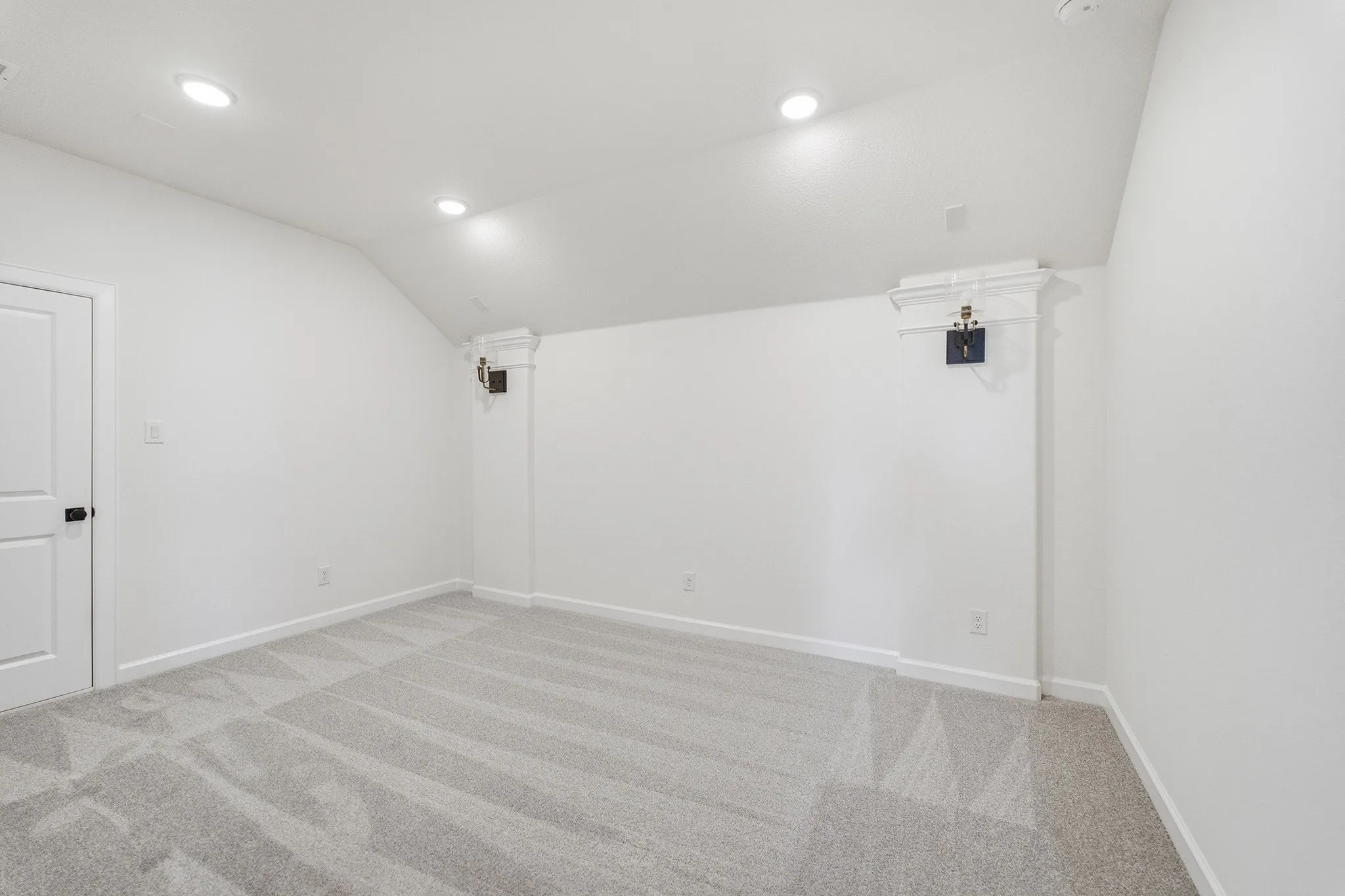 Spacious room featuring light gray patterned carpeting, recessed ceiling lighting, a white panel door with black hardware, and angled ceiling details