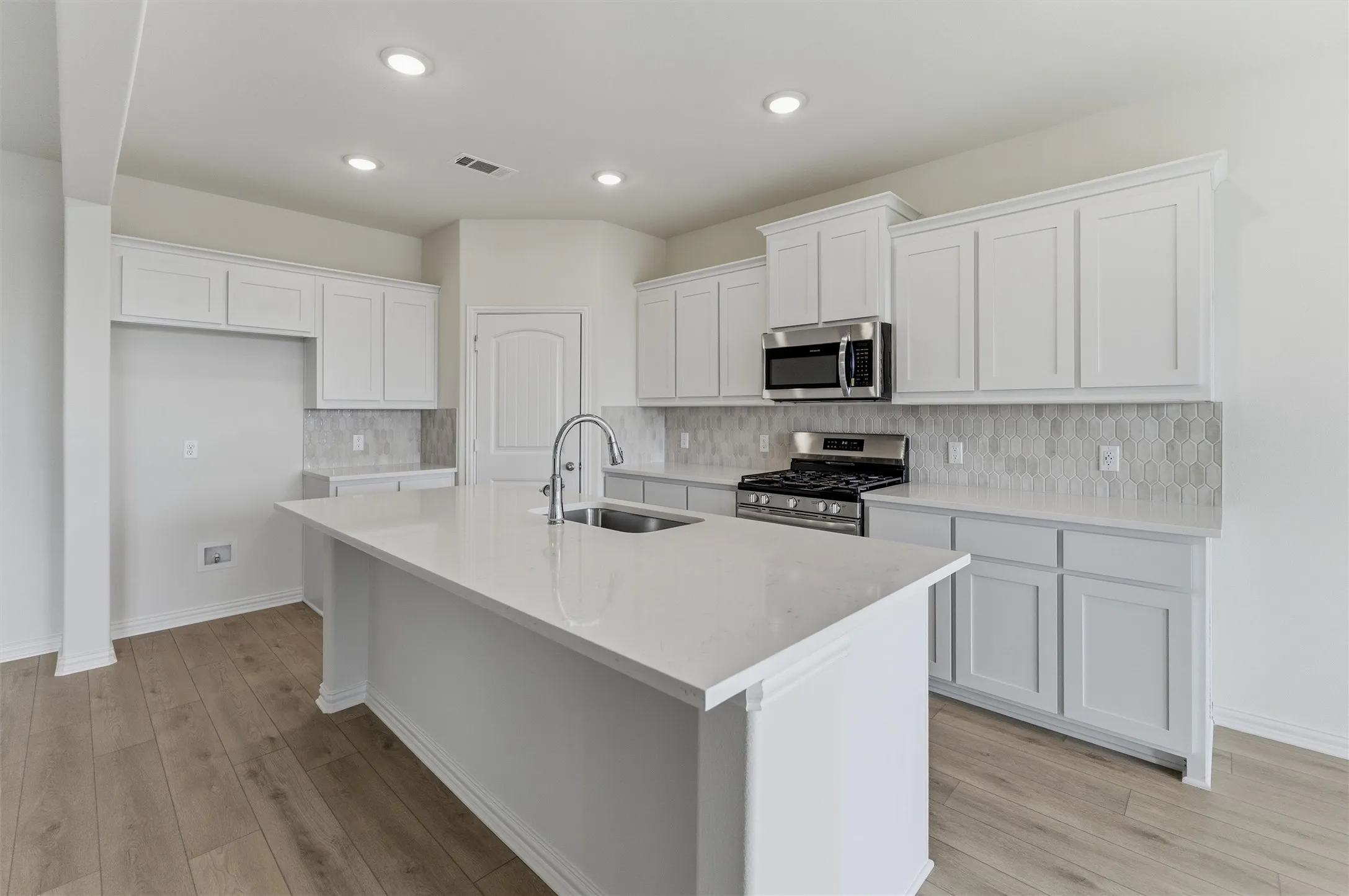 Kitchen featuring a central island with an undermount sink, stainless steel appliances, white cabinetry, a hexagonal tiled backsplash, and wood-finish flooring