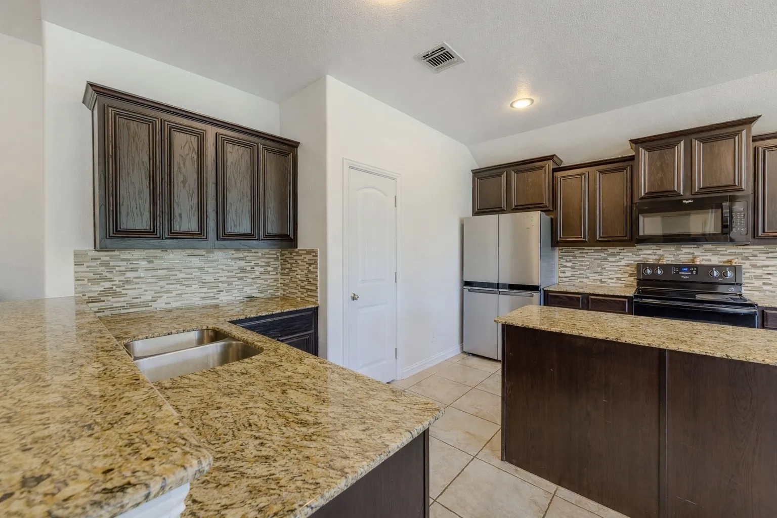 Kitchen with dark brown cabinets, black appliances, backsplash, light stone countertops, and vaulted ceiling