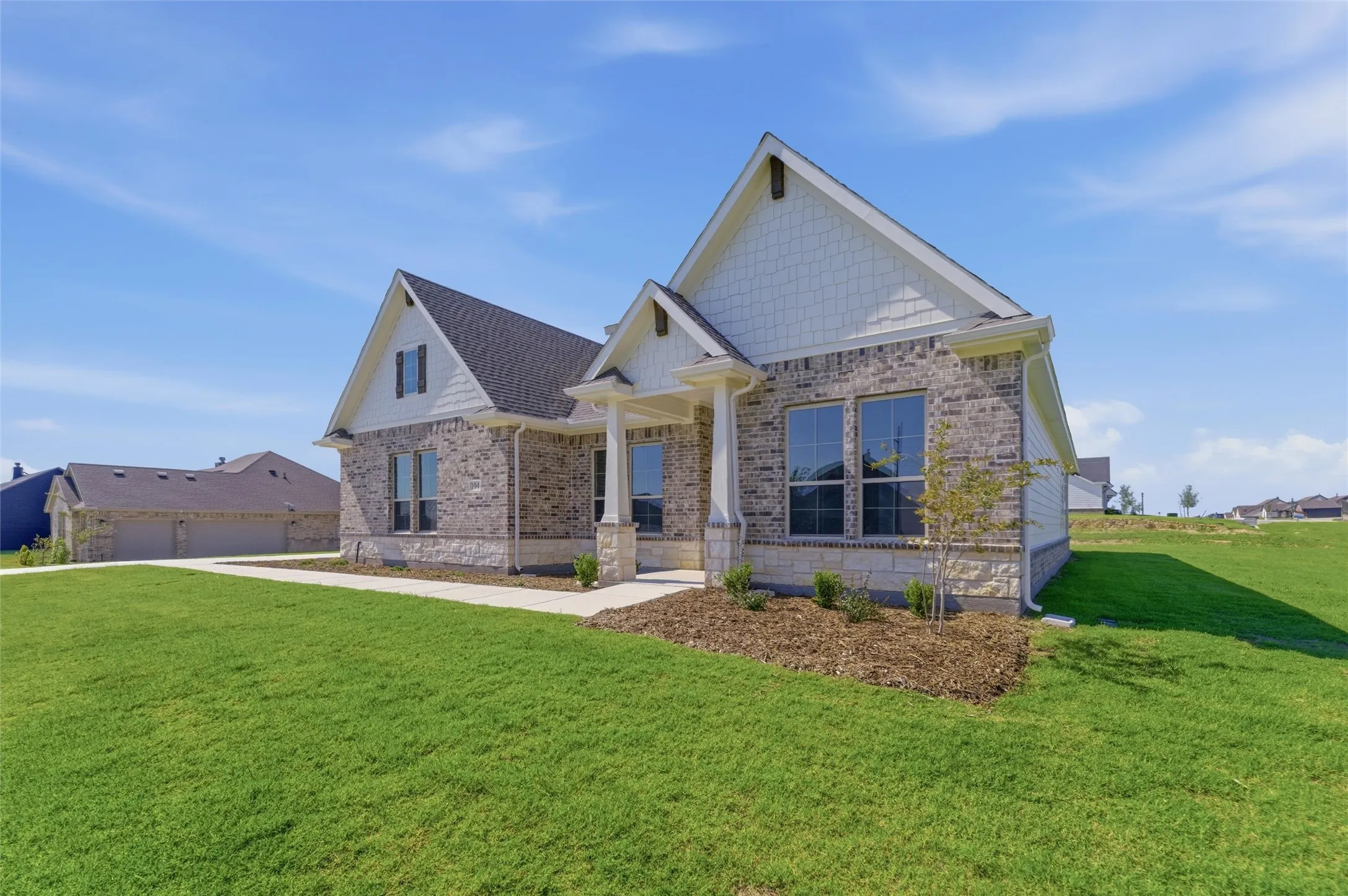 Single-story residence featuring a brick and stone facade, gabled rooflines with shingle siding accents, a covered entry with columns, and a landscaped front yard with a concrete pathway