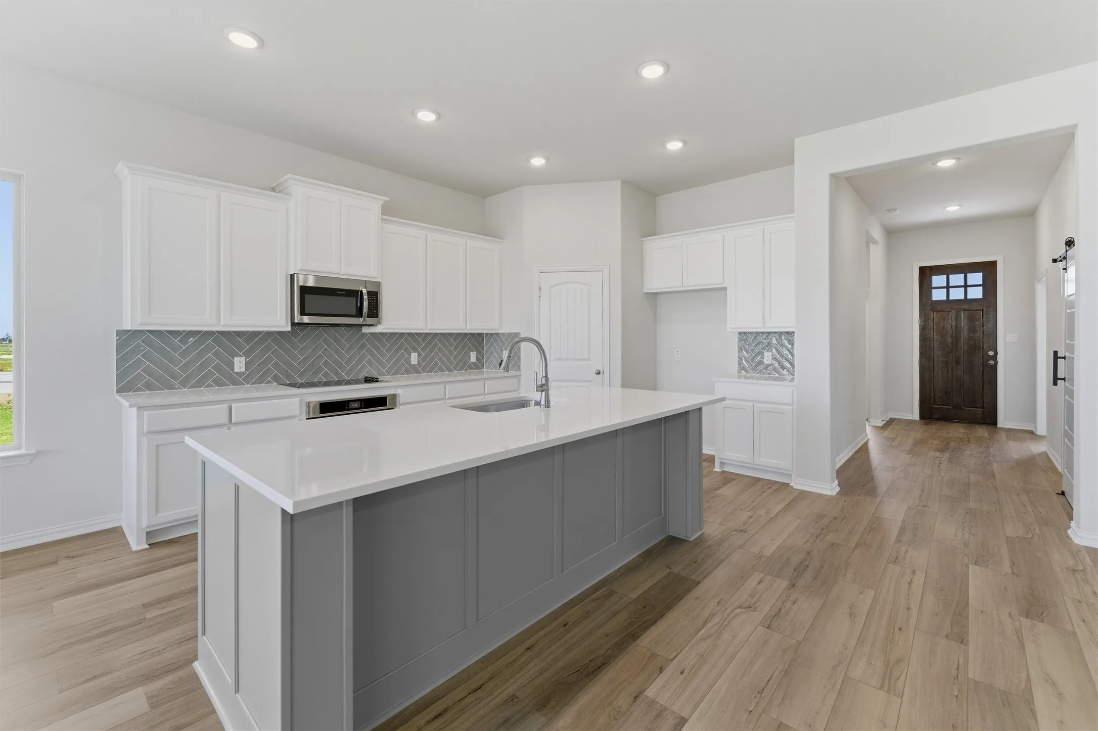 Open-concept kitchen featuring an expansive center island with a white countertop and gray paneling, stainless steel appliances, a gray herringbone tile backsplash, white shaker-style cabinetry, and wood-finish flooring