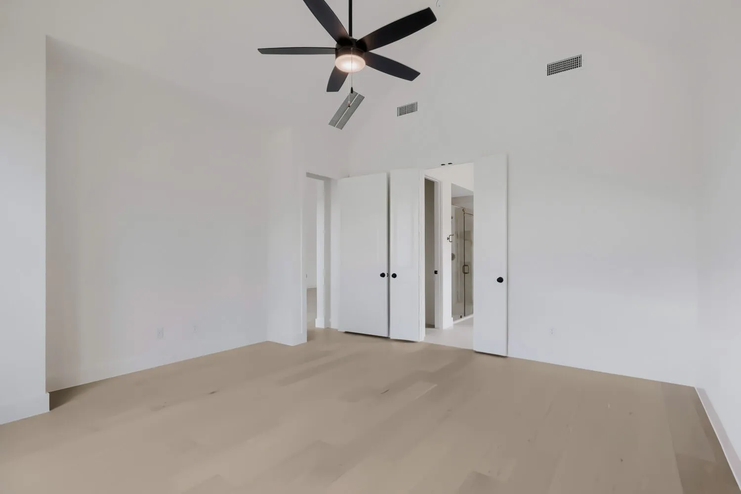 Spacious room featuring vaulted ceilings, a black ceiling fan with integrated lighting, light wood-finish flooring, and white bifold doors revealing an en-suite bathroom with a glass-enclosed shower