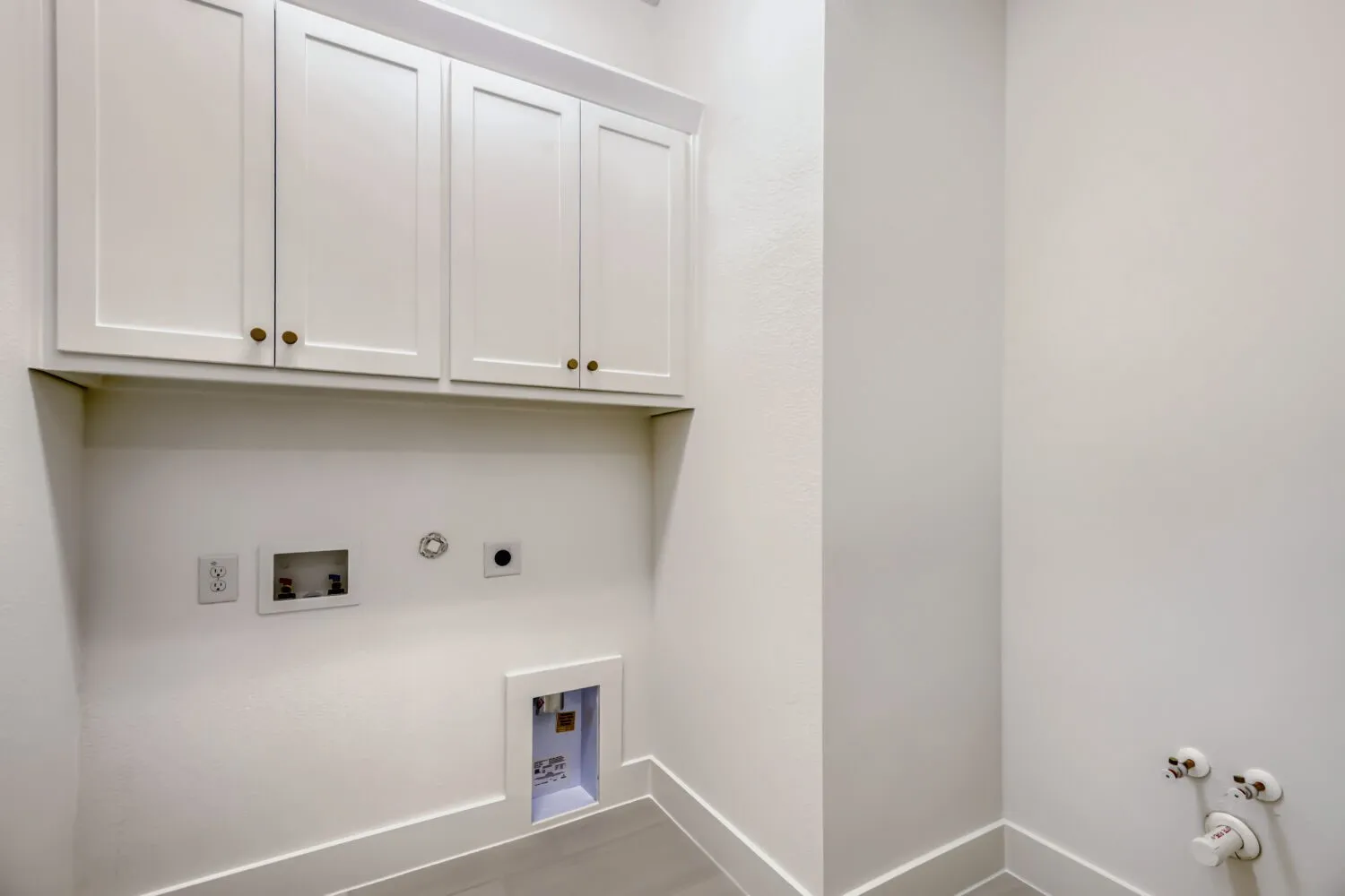 Laundry area featuring shaker-style upper cabinetry with brass-toned hardware, white walls, and light-toned tile flooring