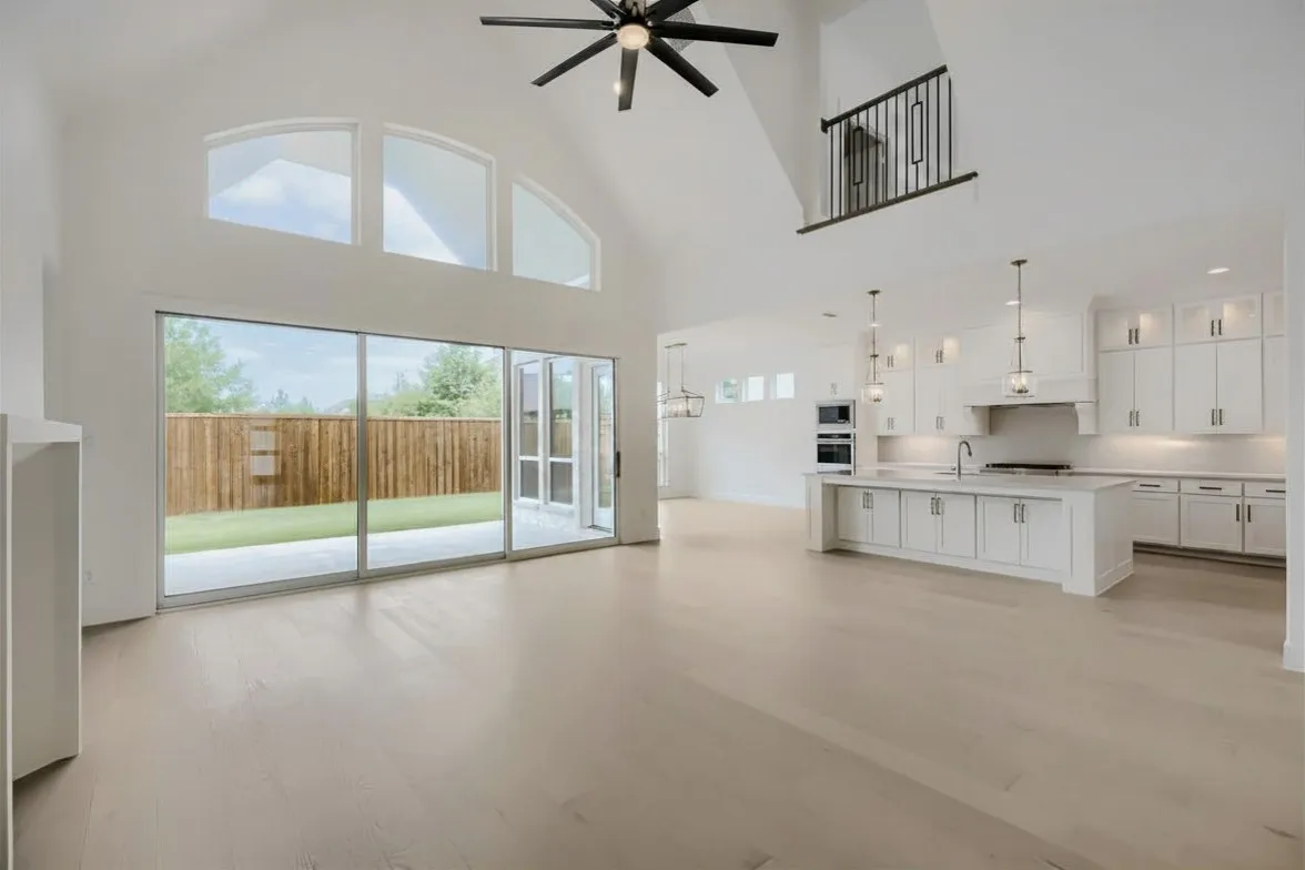 Vaulted ceiling great room featuring wood-finish flooring, a contemporary kitchen with white cabinetry and an island, a modern ceiling fan, and extensive sliding glass doors opening to a fenced backyard