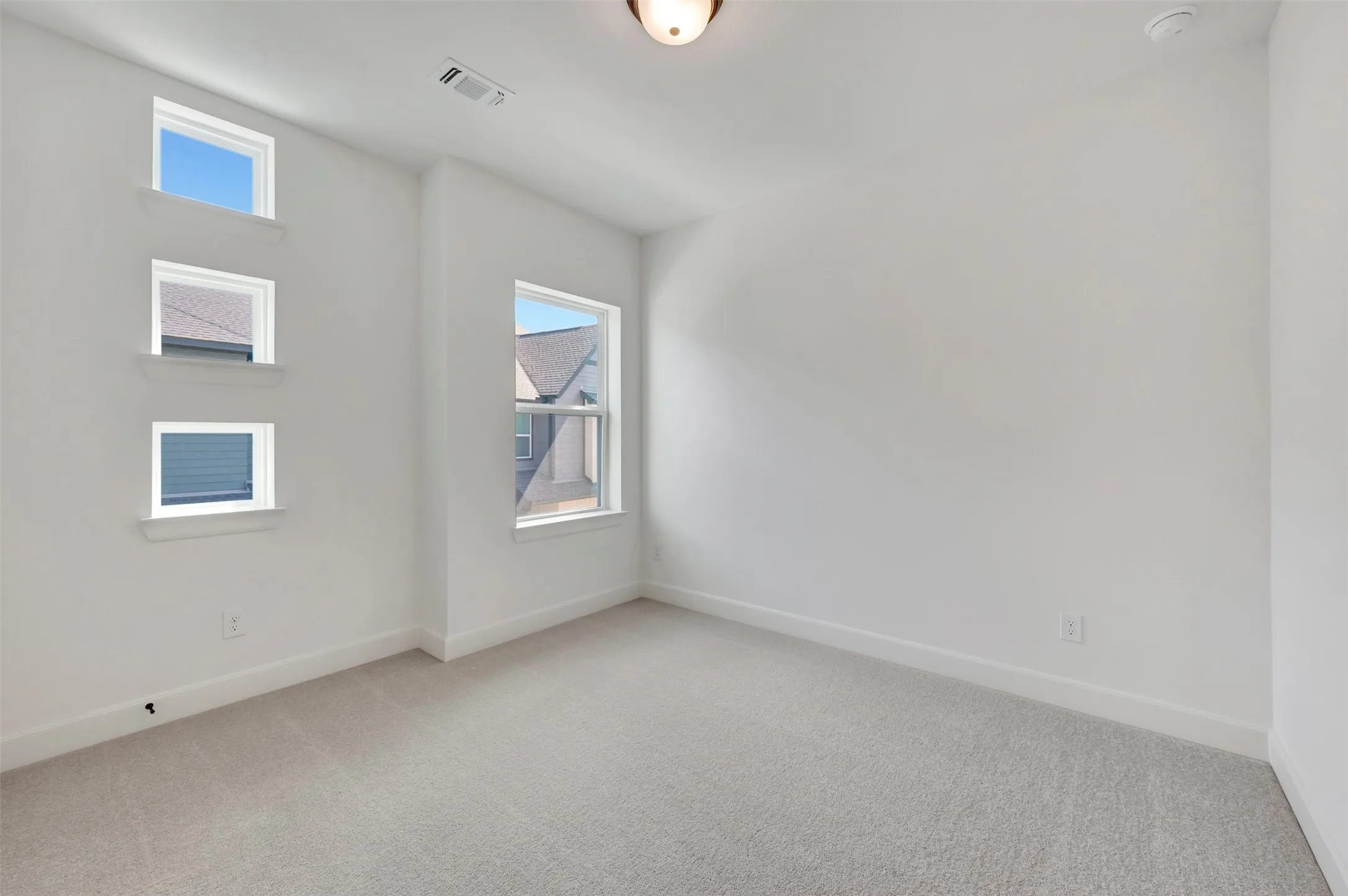 Bright room featuring three stacked windows and one rectangular window, light-toned carpeting, white walls, and a ceiling-mounted light fixture