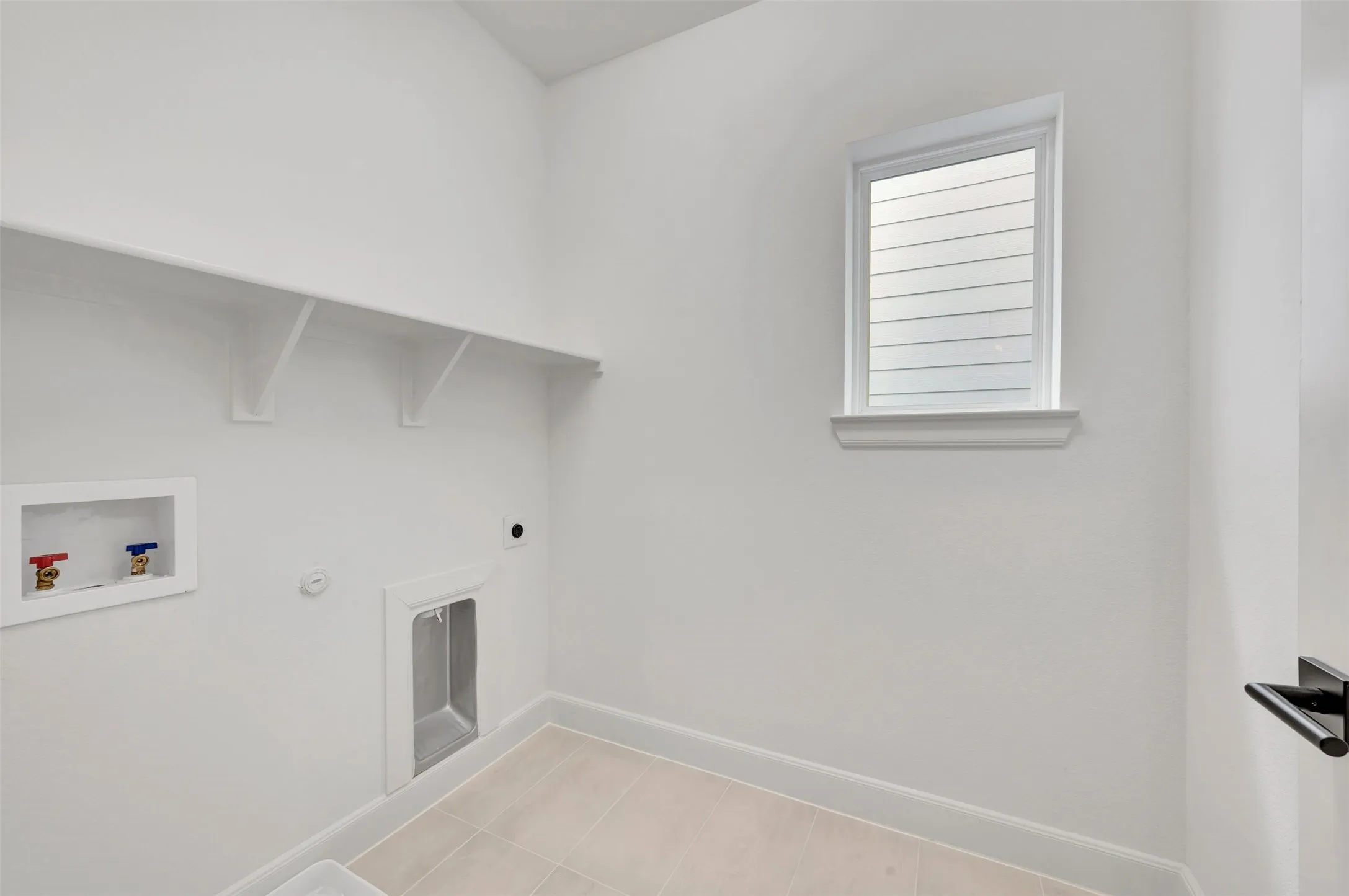 Dedicated laundry space featuring a window with integrated blinds, a wall-mounted shelf, and light-toned tile flooring
