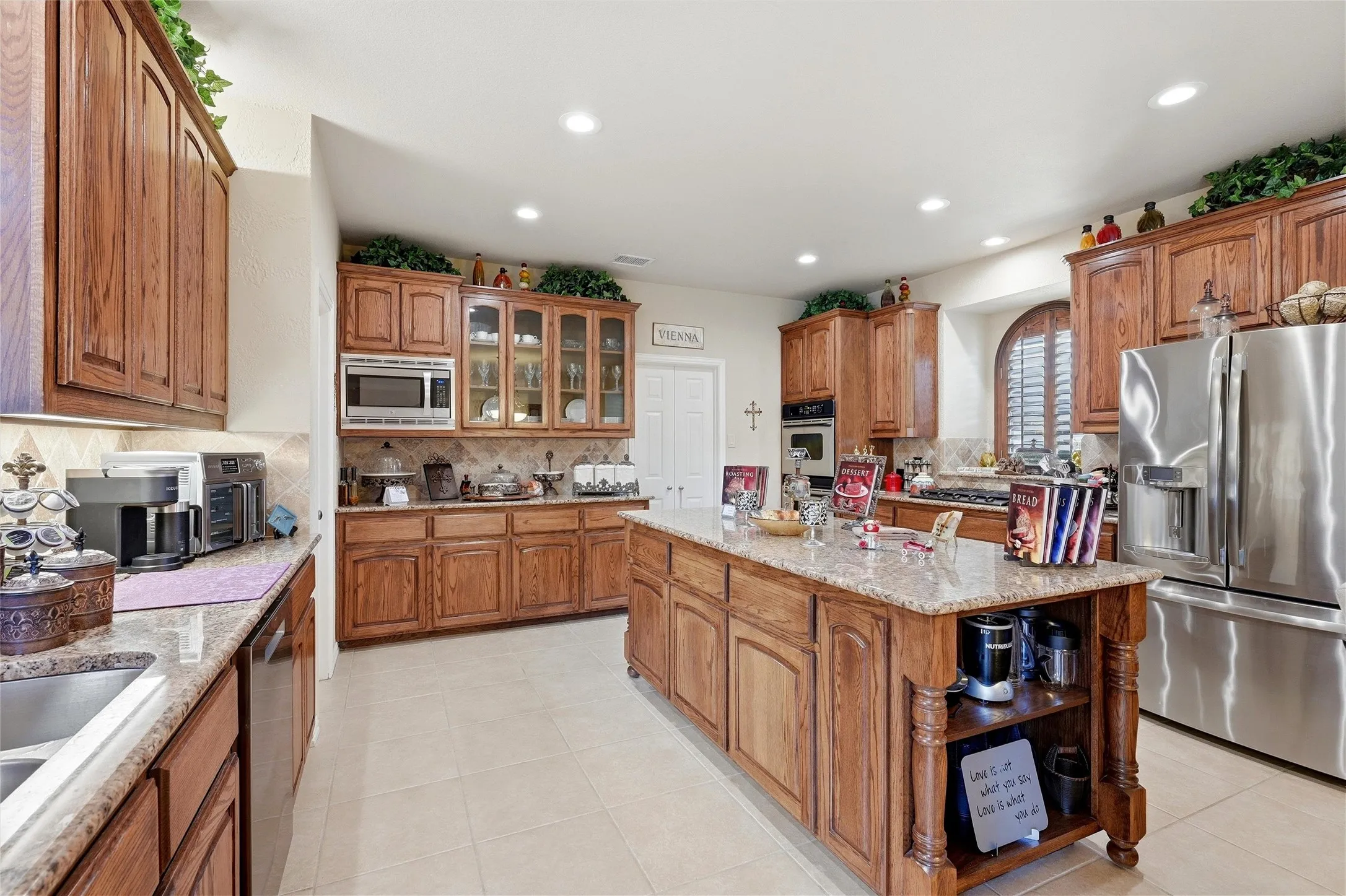 Kitchen featuring appliances with stainless steel finishes, glass insert cabinets, light stone counters, brown cabinetry, and recessed lighting