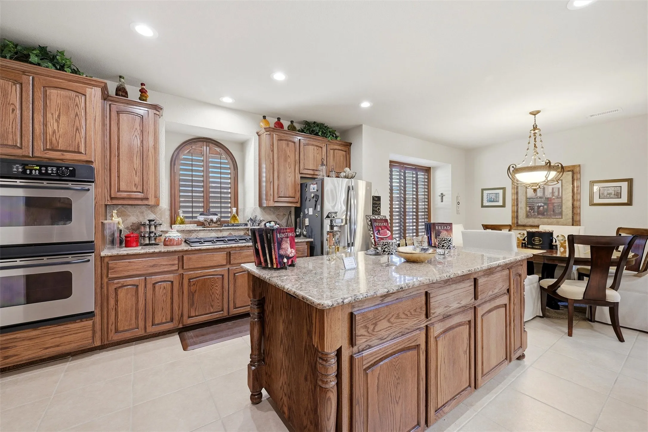 Kitchen with brown cabinetry, appliances with stainless steel finishes, light stone countertops, hanging light fixtures, and a kitchen island