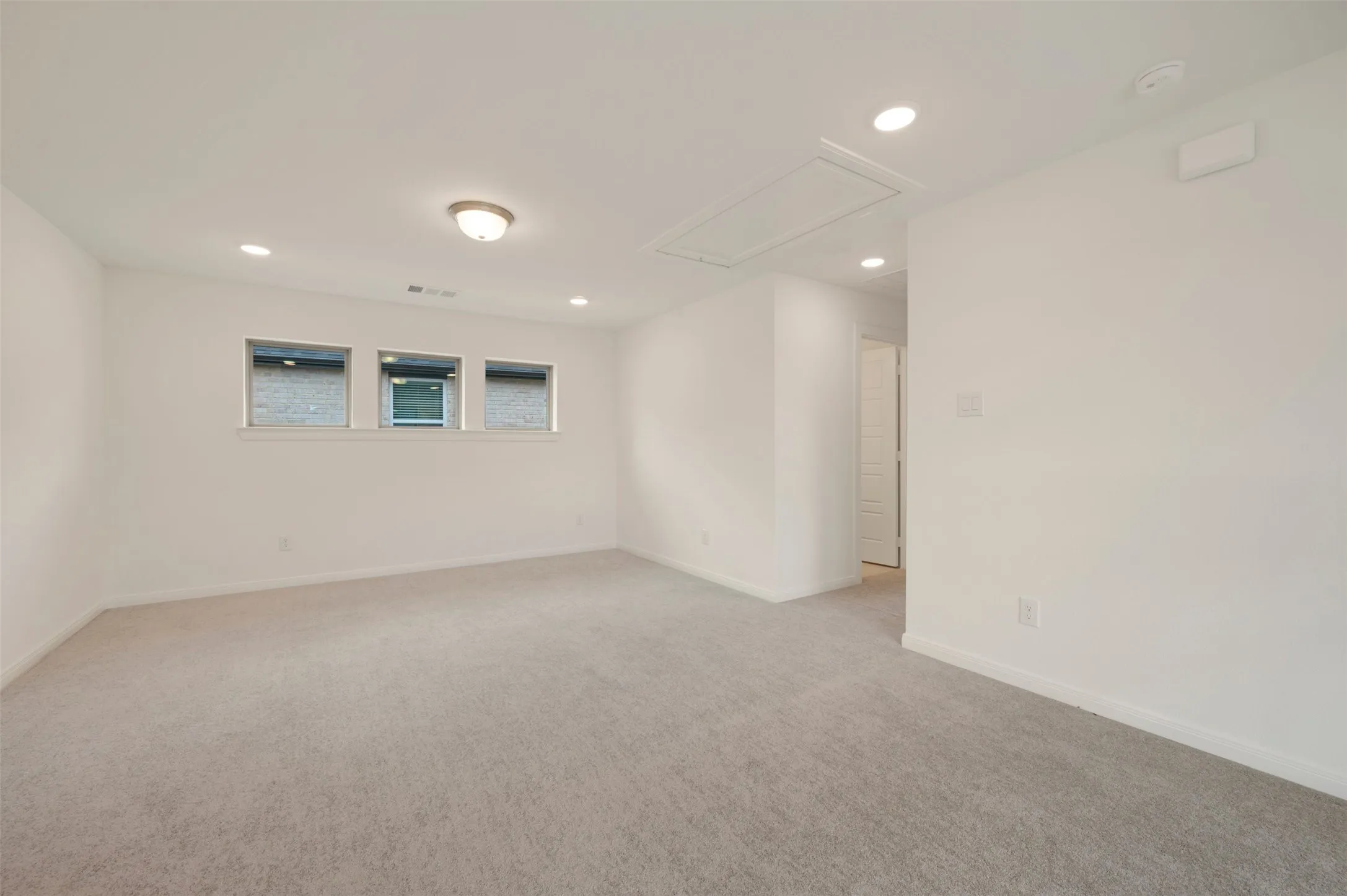 Spacious room featuring neutral carpeting, three horizontal windows with white trim, and a combination of recessed lighting and a central flush-mount fixture
