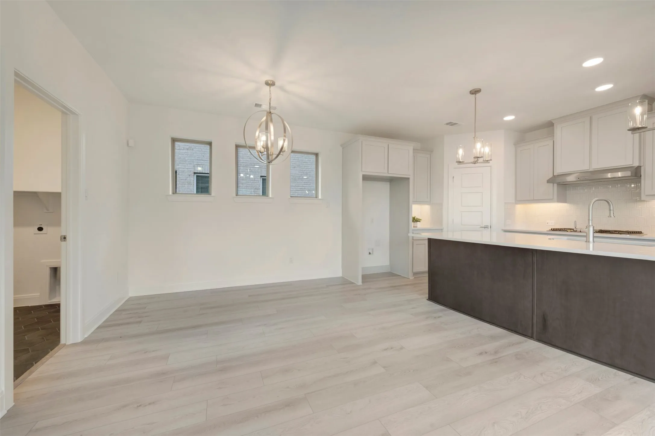 Open-concept area featuring light wood-finish flooring, a contemporary spherical chandelier, three rectangular windows, a kitchen island with a light-toned countertop, and white cabinetry