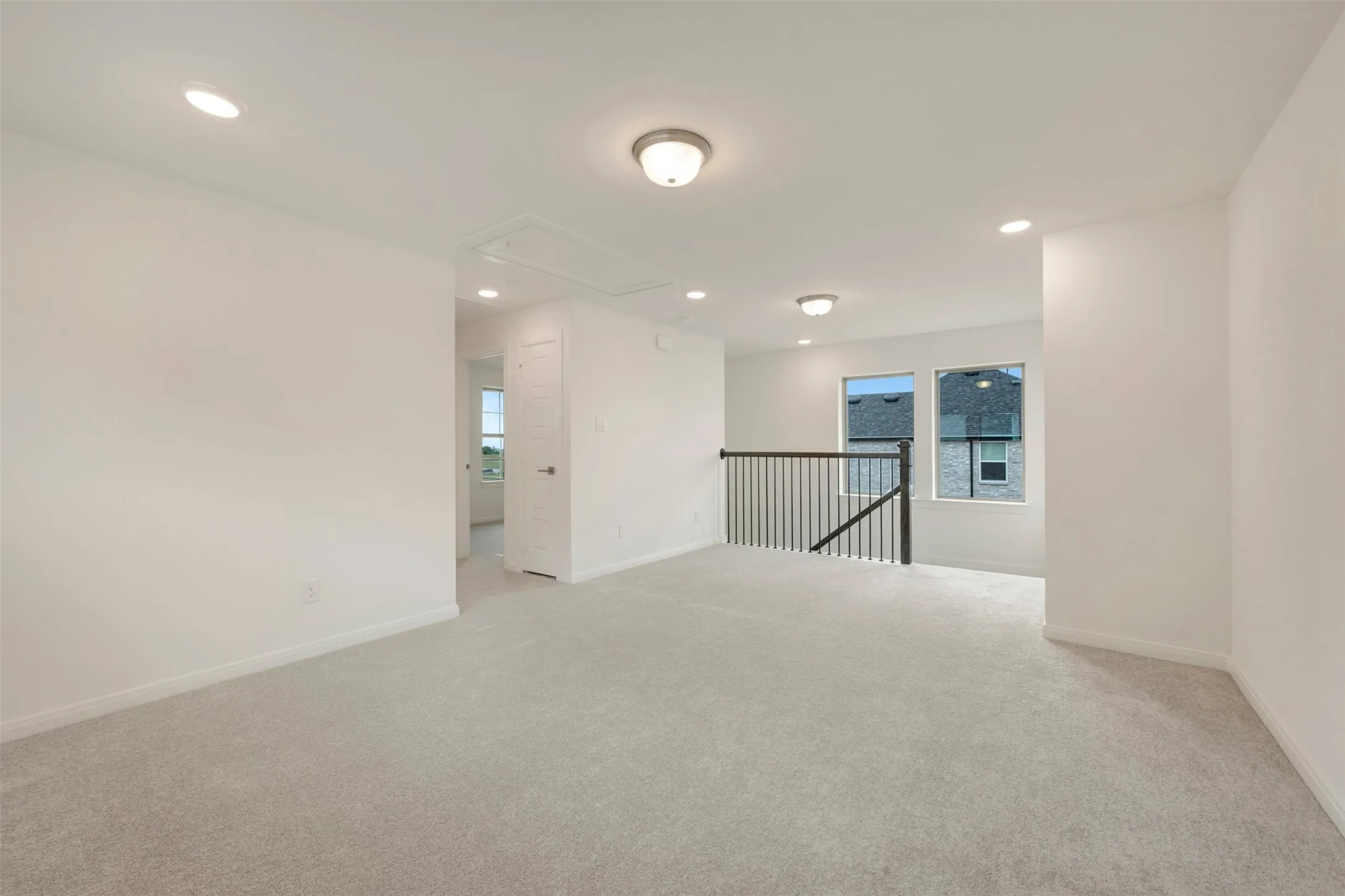 Spacious secondary living area featuring light gray carpeting, recessed lighting, and a dark wood-finish staircase railing with black spindles