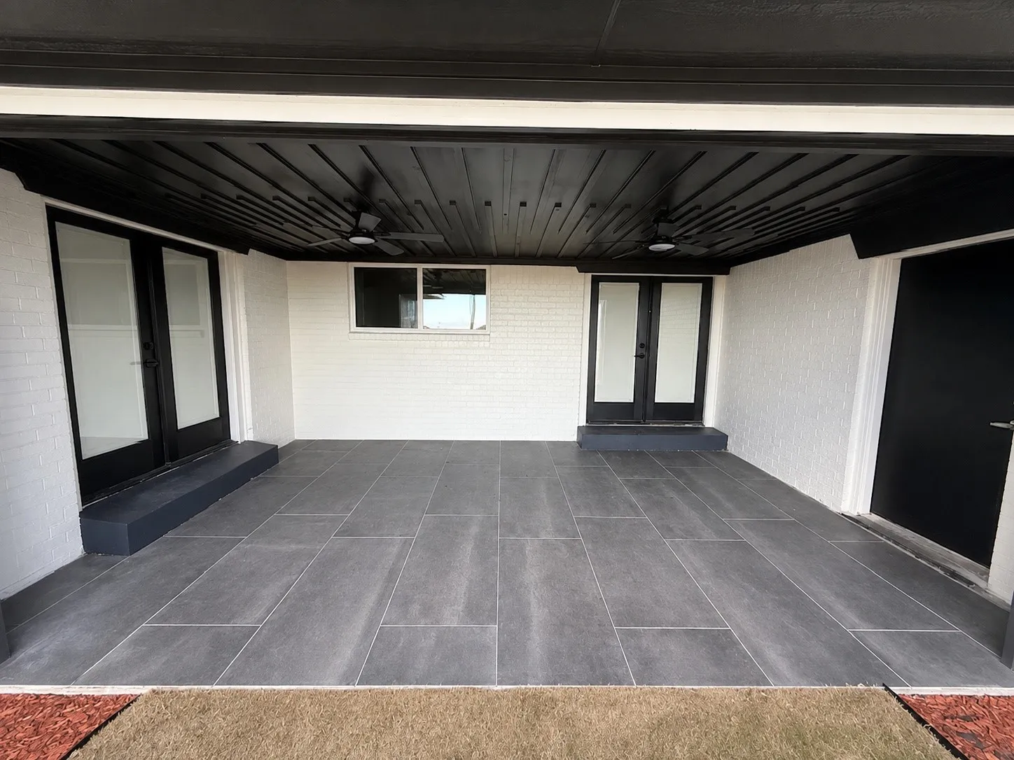 Covered patio featuring large format grey tile flooring, white brick walls, and a black wood-finish ceiling with recessed lighting