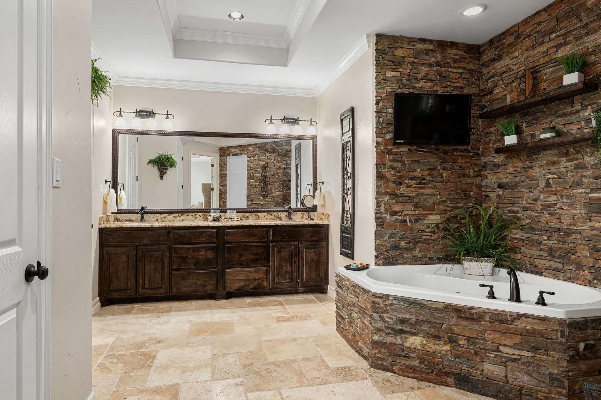 Full bath featuring double vanity, stone tile flooring, a garden tub, crown molding, and a raised ceiling